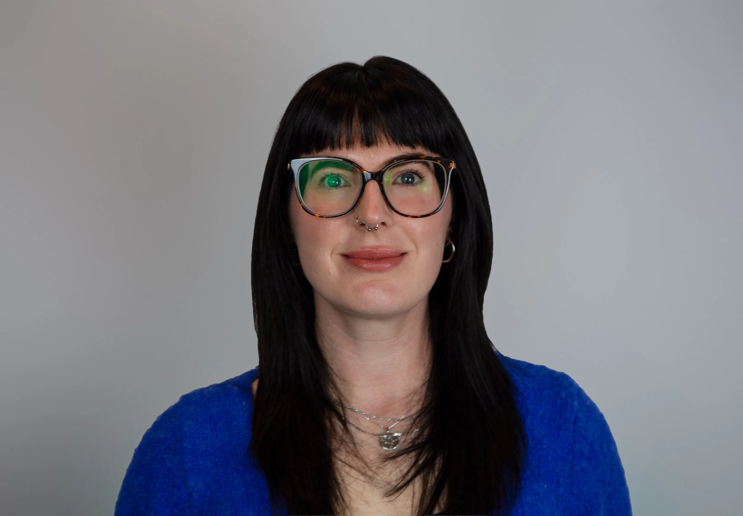 Portrait of a woman with dark hair, wearing large glasses and a blue sweater, smiling slightly against a plain gray background.