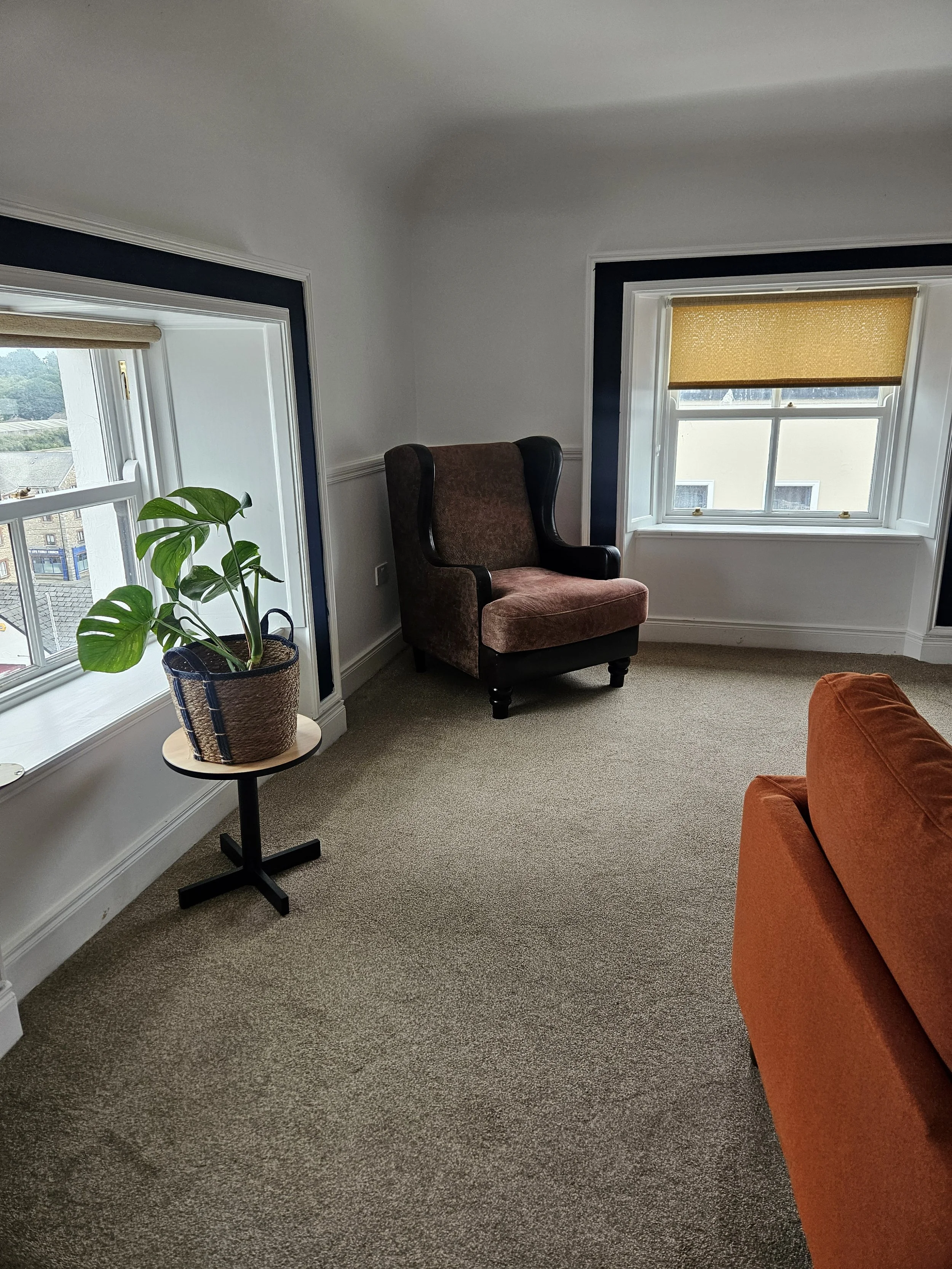A cozy corner of a room with beige carpeted floor, a brown upholstered armchair, a small round side table with a potted plant, and two windows with white trim and yellow blinds, allowing natural light.