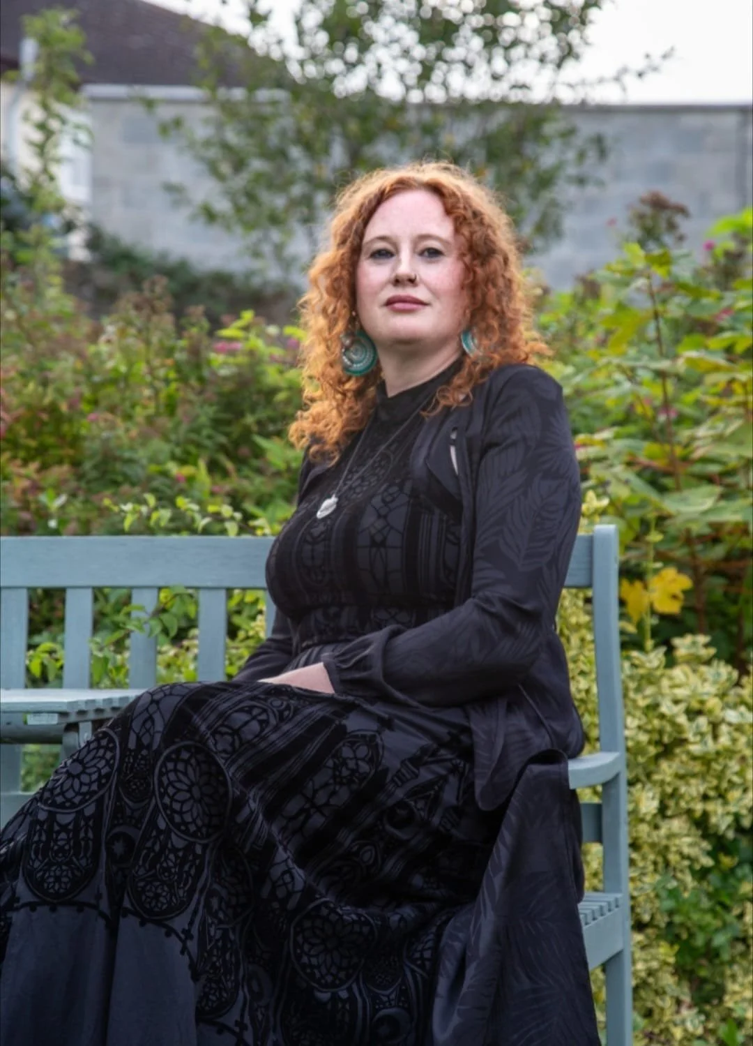 A woman with curly red hair sitting on a blue bench outdoors, surrounded by green and yellow foliage, wearing a black dress and earrings.
