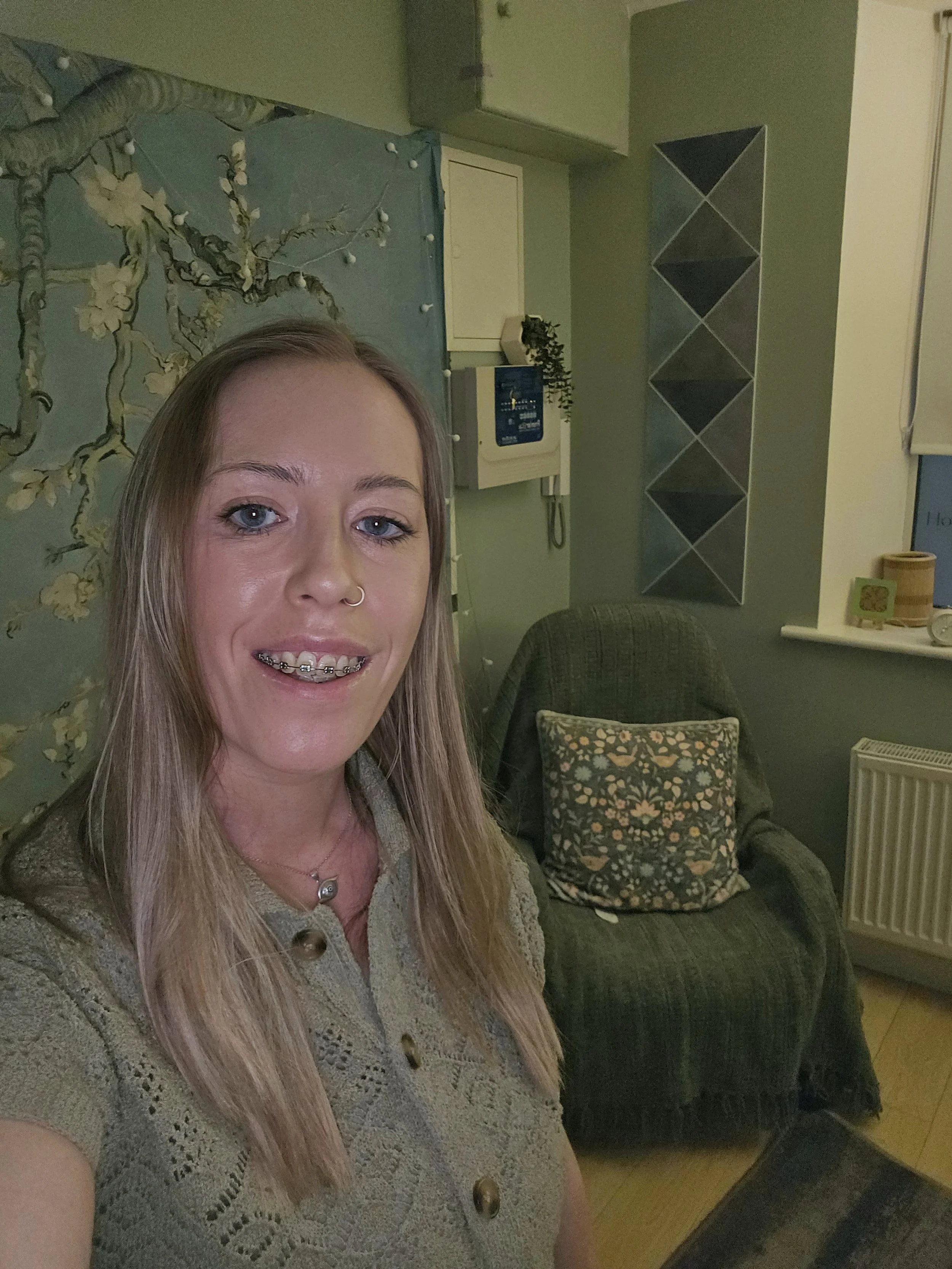 A young woman with long blond hair, braces, and a nose ring taking a selfie in a room with green walls, a patterned armchair, and decorative wall art.