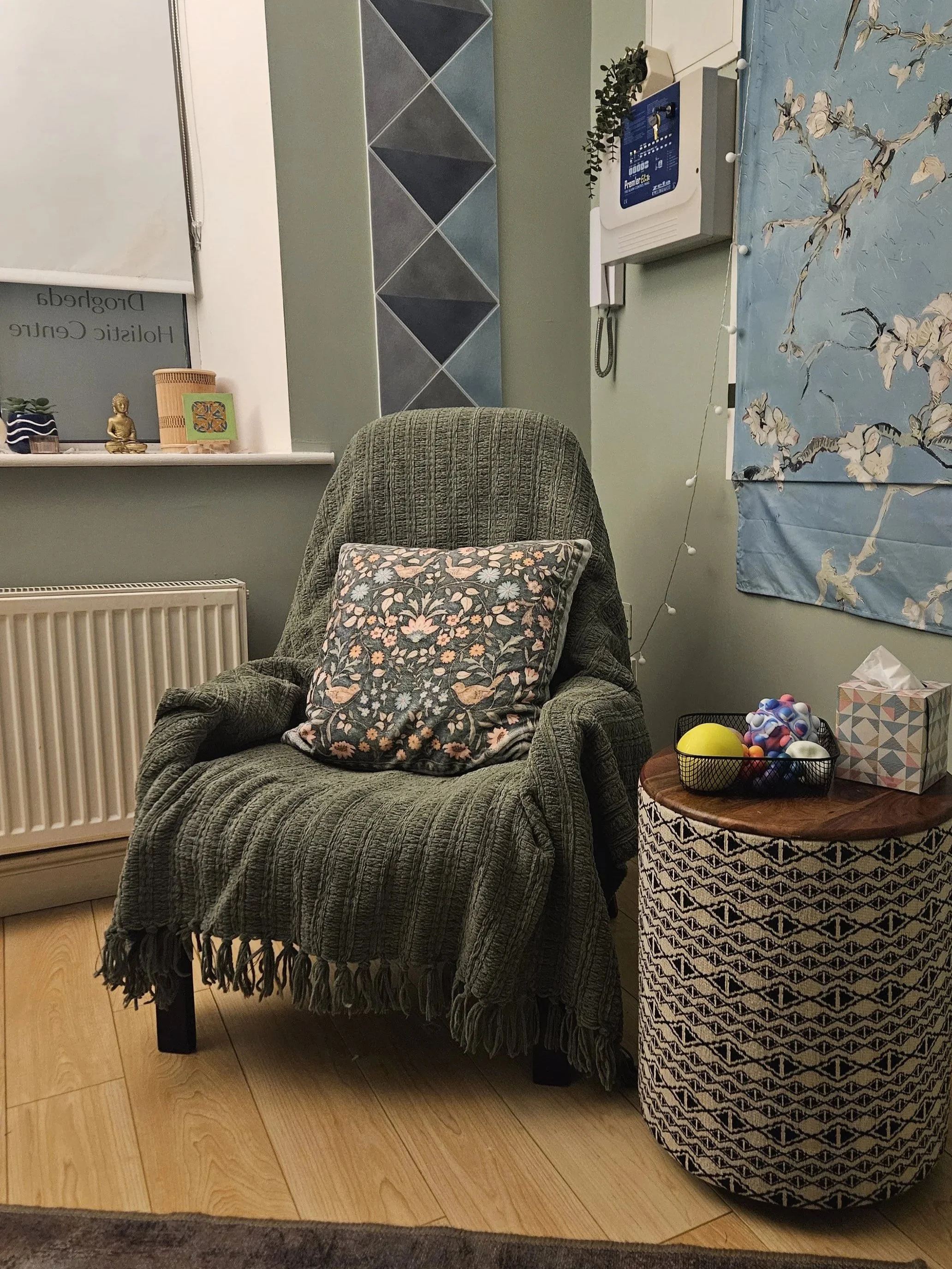 A cozy chair with a floral pillow, next to a woven side table with a ball of yarn and a tissue box, in a room with a window, radiator, and wall decorations.