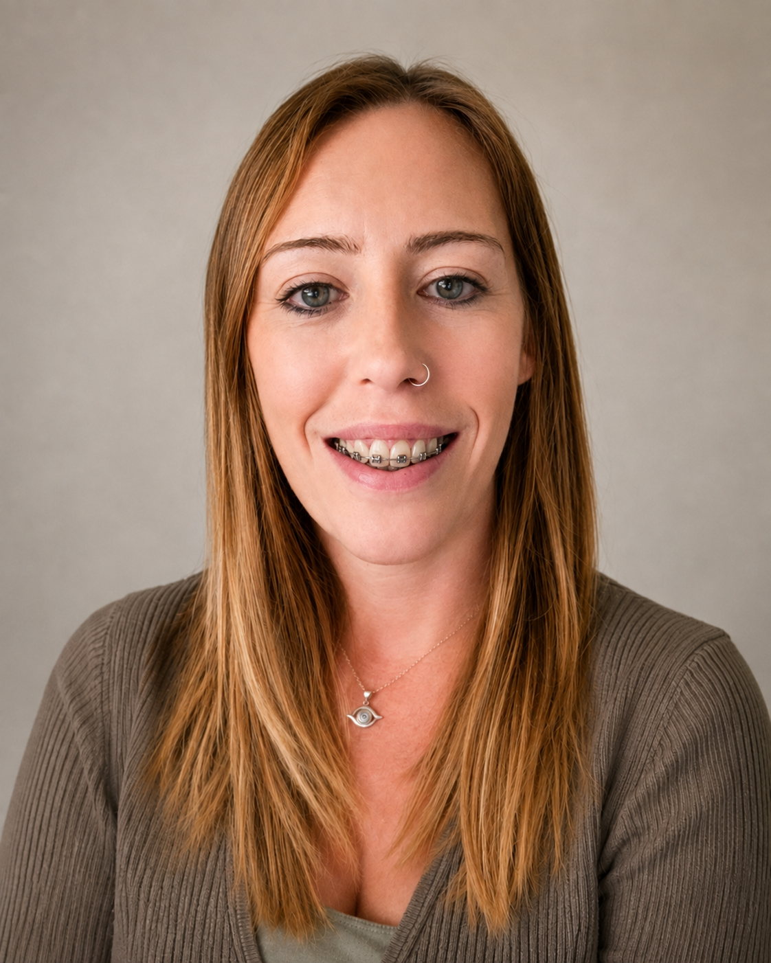 A woman with long red hair, wearing a brown ribbed top and a necklace with an eye pendant, smiling with braces showing, against a neutral background.