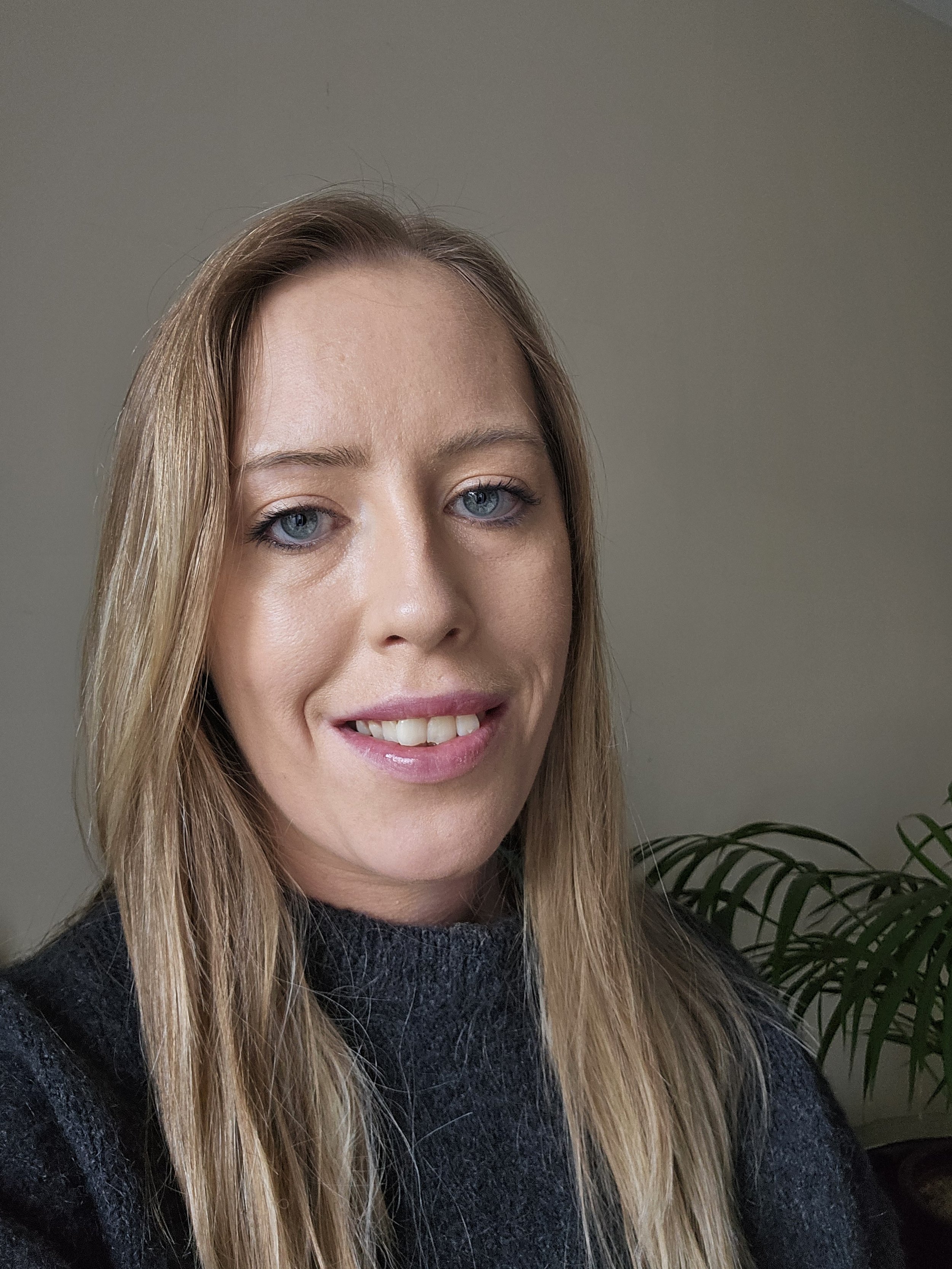 A woman with blonde hair and blue eyes smiling and wearing a dark sweater, standing indoors near a plant.