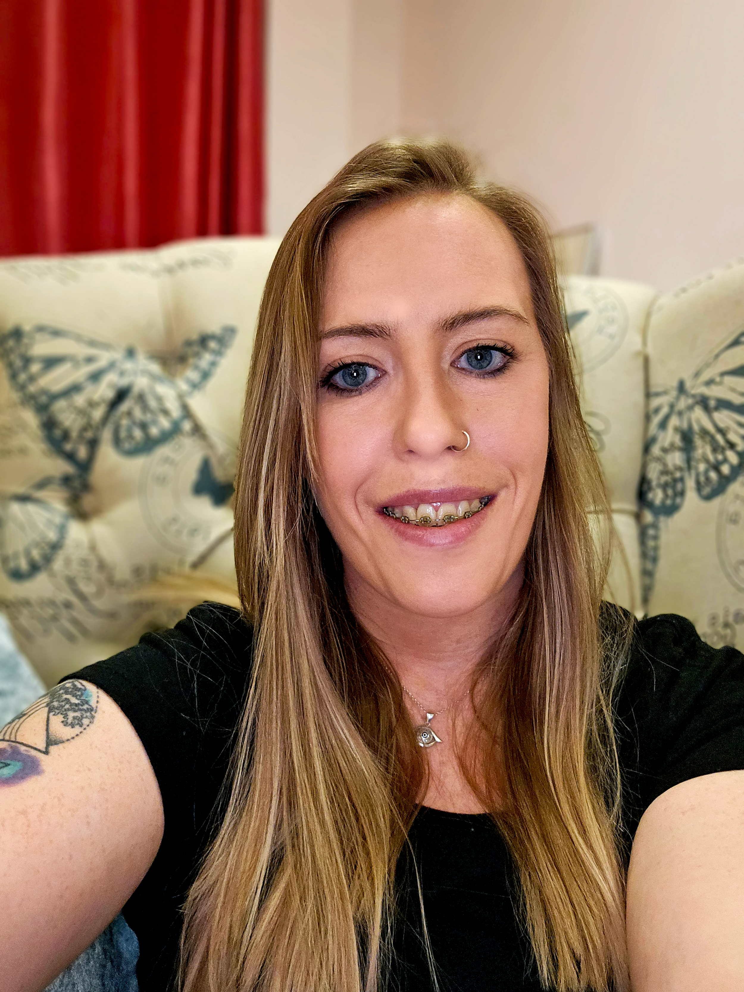 A woman with long blonde hair, blue eyes, wearing a black shirt, and a nose ring, smiling for a selfie in a room with butterfly patterned pillows and red curtains.
