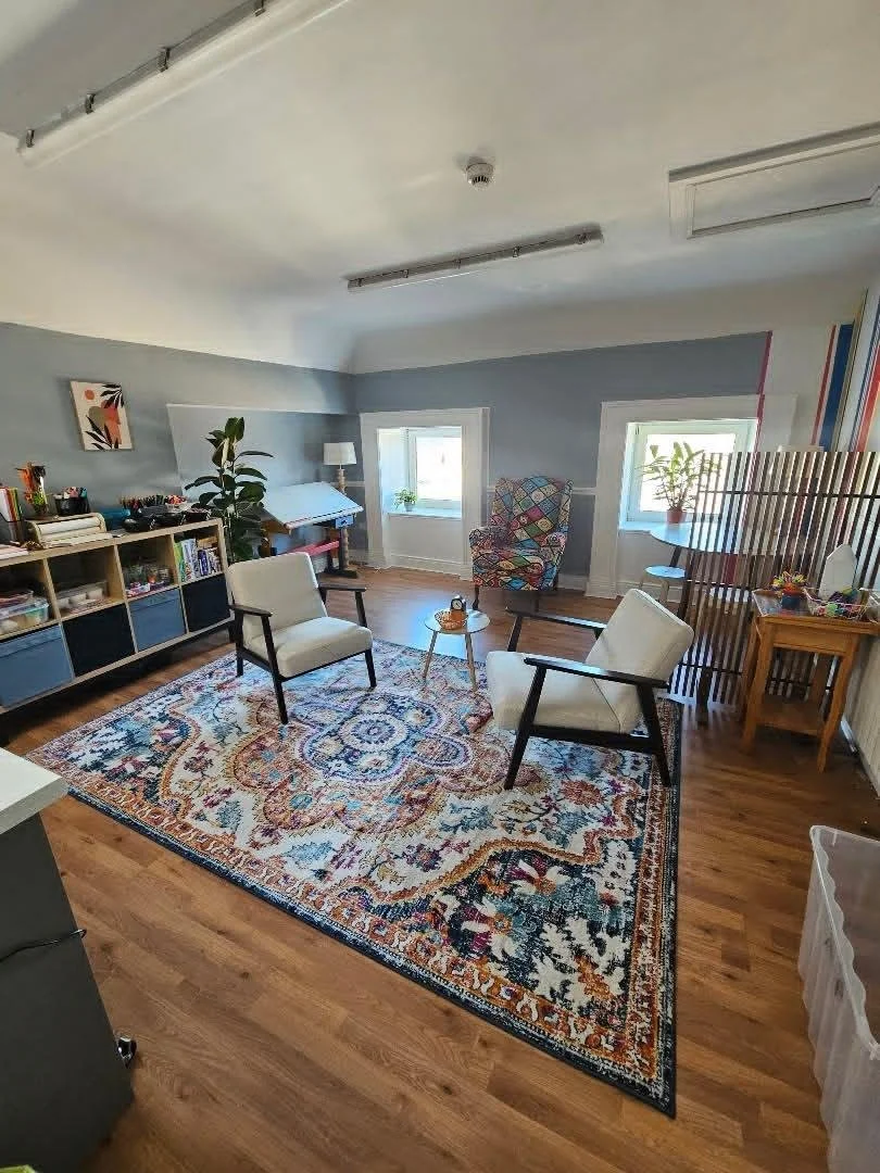 A cozy living room with a wooden floor, a patterned area rug, and two white armchairs with black frames. There are two small round tables, a patterned armchair, and plants near the windows. A bookshelf with various items and books is along the back wall.