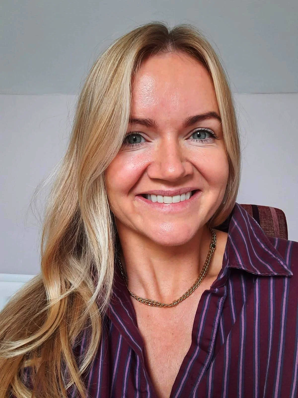 Close-up of a smiling blonde woman with blue eyes, wearing a striped purple shirt and gold necklace, sitting on a chair with a plain light-colored wall in the background.