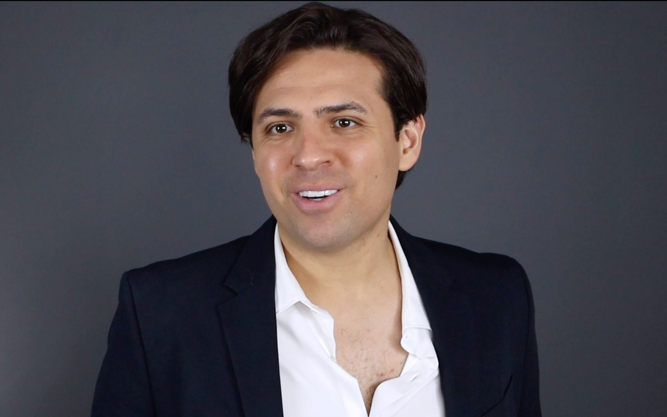 Portrait of a man with dark hair, wearing a black blazer over a white shirt, smiling against a gray background.