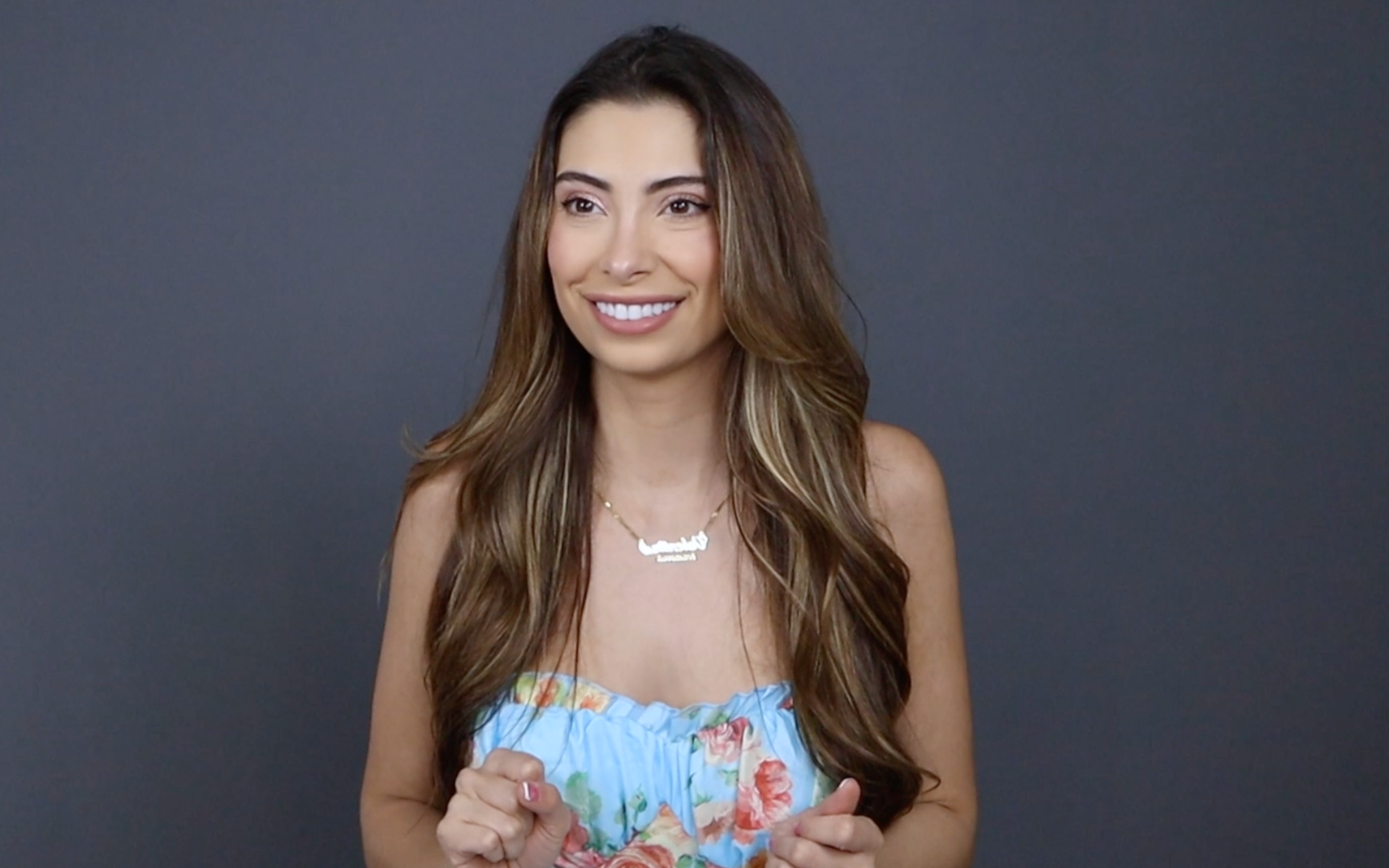 Smiling woman with long brown hair wearing a light blue floral dress and a necklace, in front of a gray background.