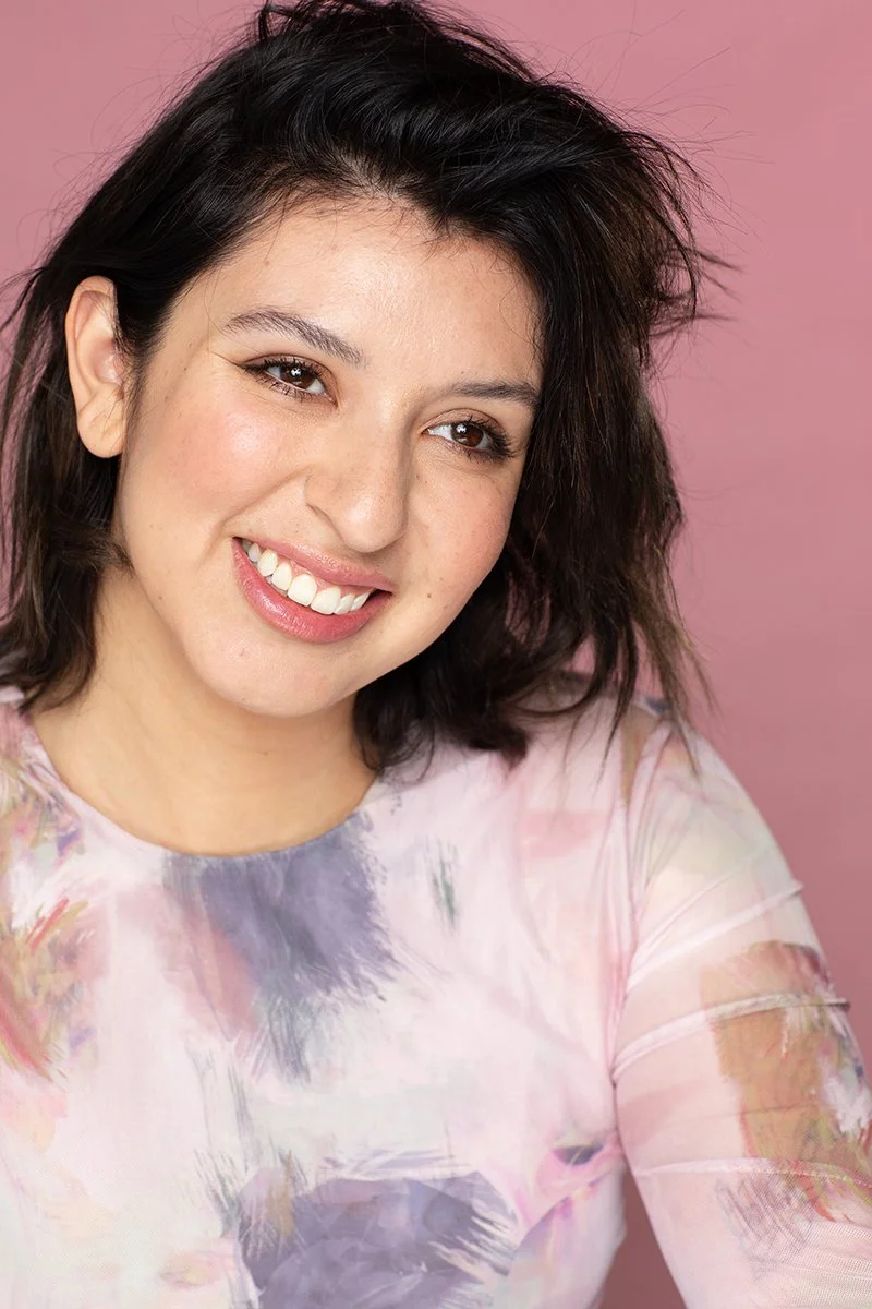 A woman with dark, wavy hair smiling in front of a pink background, wearing a pastel tie-dye shirt.