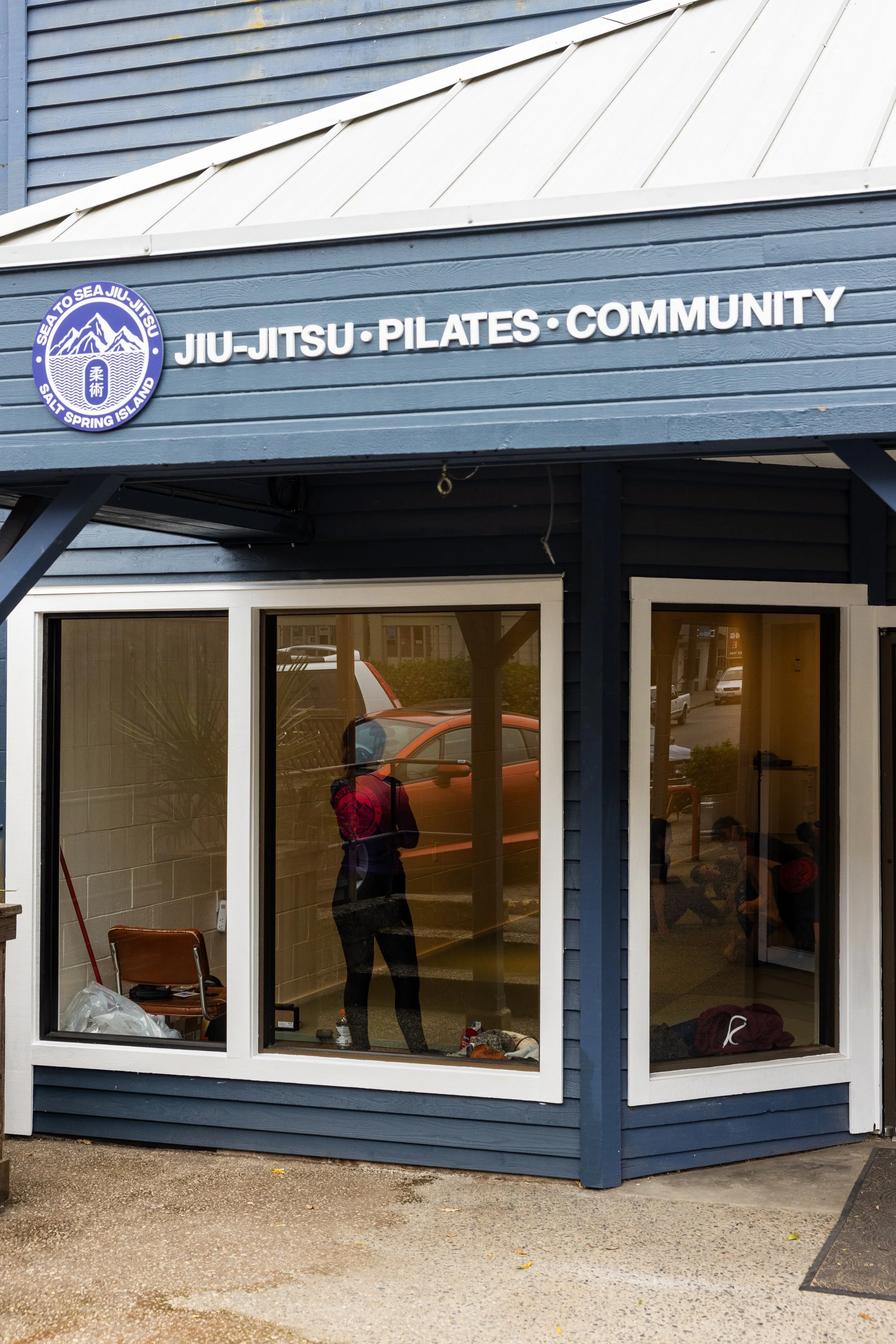 Exterior of a building with a large window and a sign that reads 'Jiu-Jitsu, Pilates, Community'. The building's siding is painted dark blue, and the window reflects parked cars and the street outside. Inside, there are chairs and various items visible through the glass.