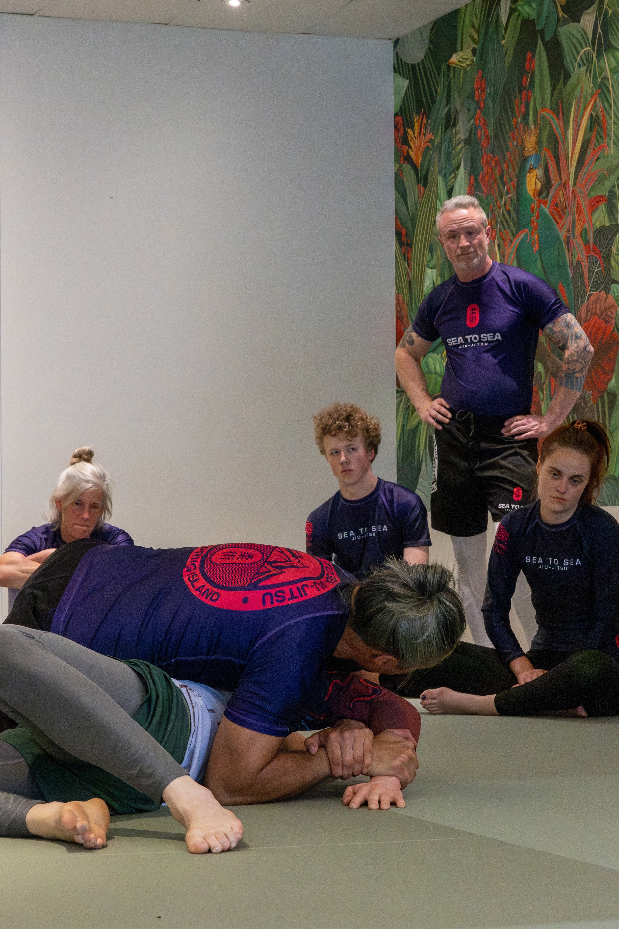 People participating in a martial arts training session, with some performing a plank and others standing watch, in a room with a tropical-themed mural.