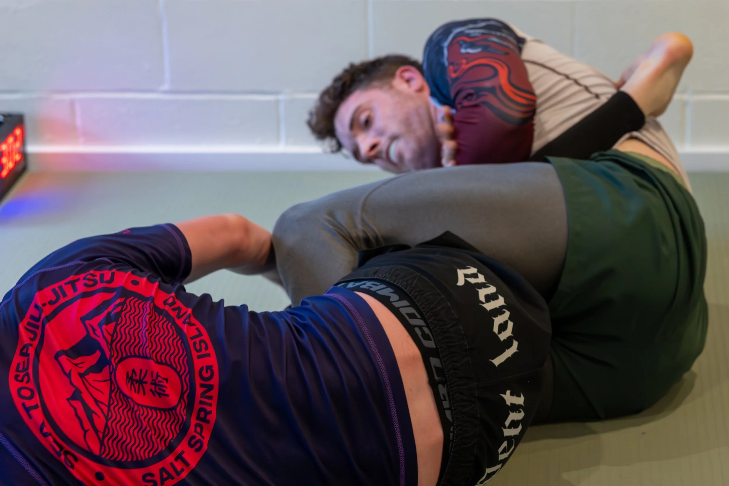 A man in athletic clothing, including shorts and a shirt, is lying on a padded floor next to a wall with an elevator button, appearing to be in a wrestling or martial arts training session.