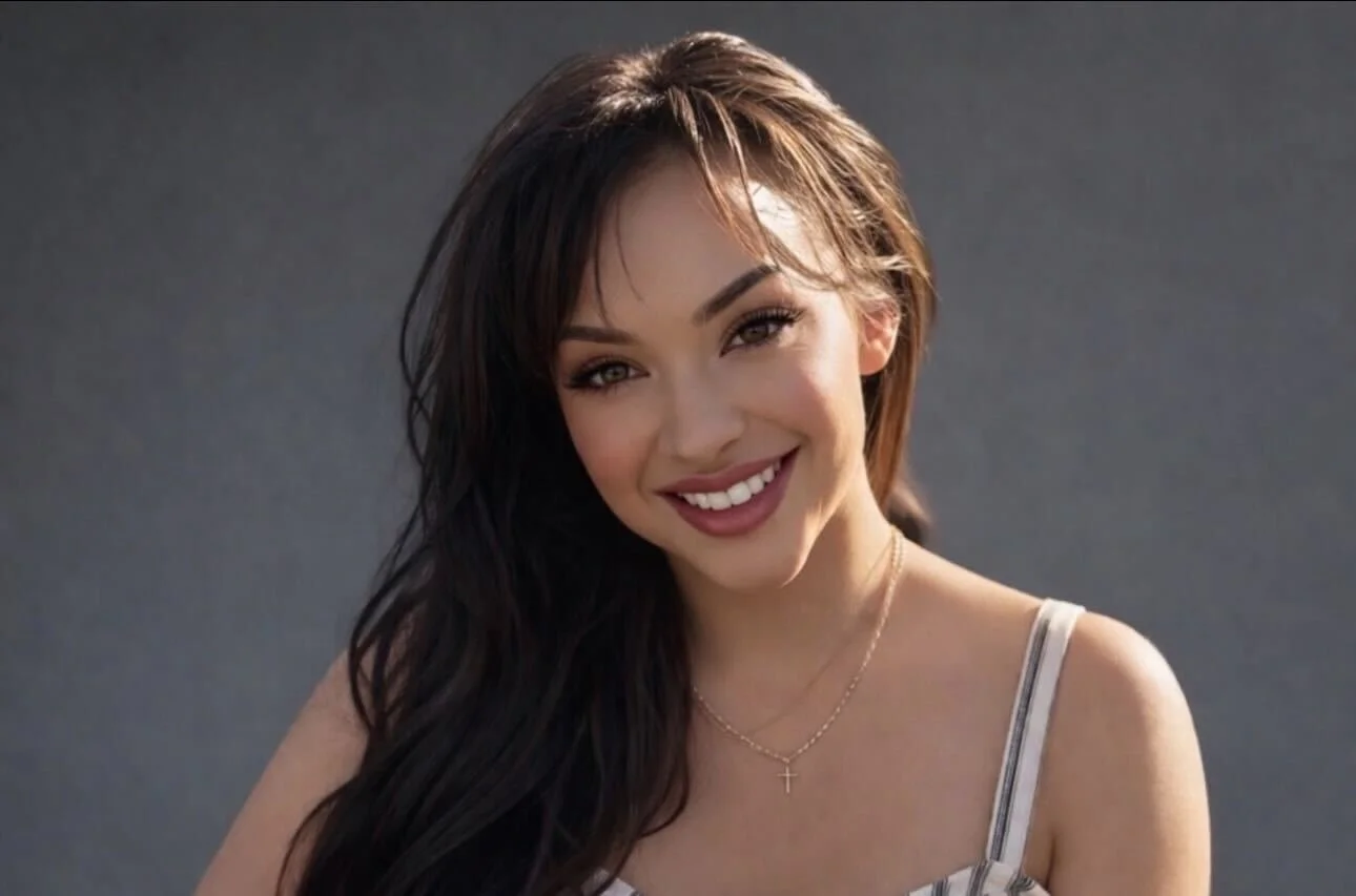 A smiling young woman with long dark hair, wearing a striped tank top and a delicate cross necklace, posing against a plain dark background.