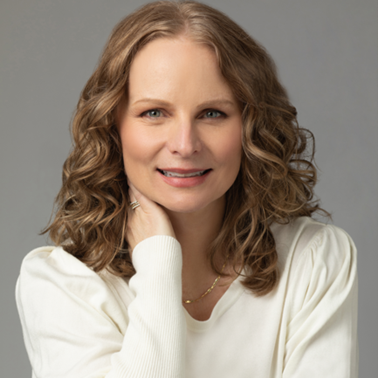 Woman with curly brown hair smiling and wearing a white sweater, posing against a plain gray background.
