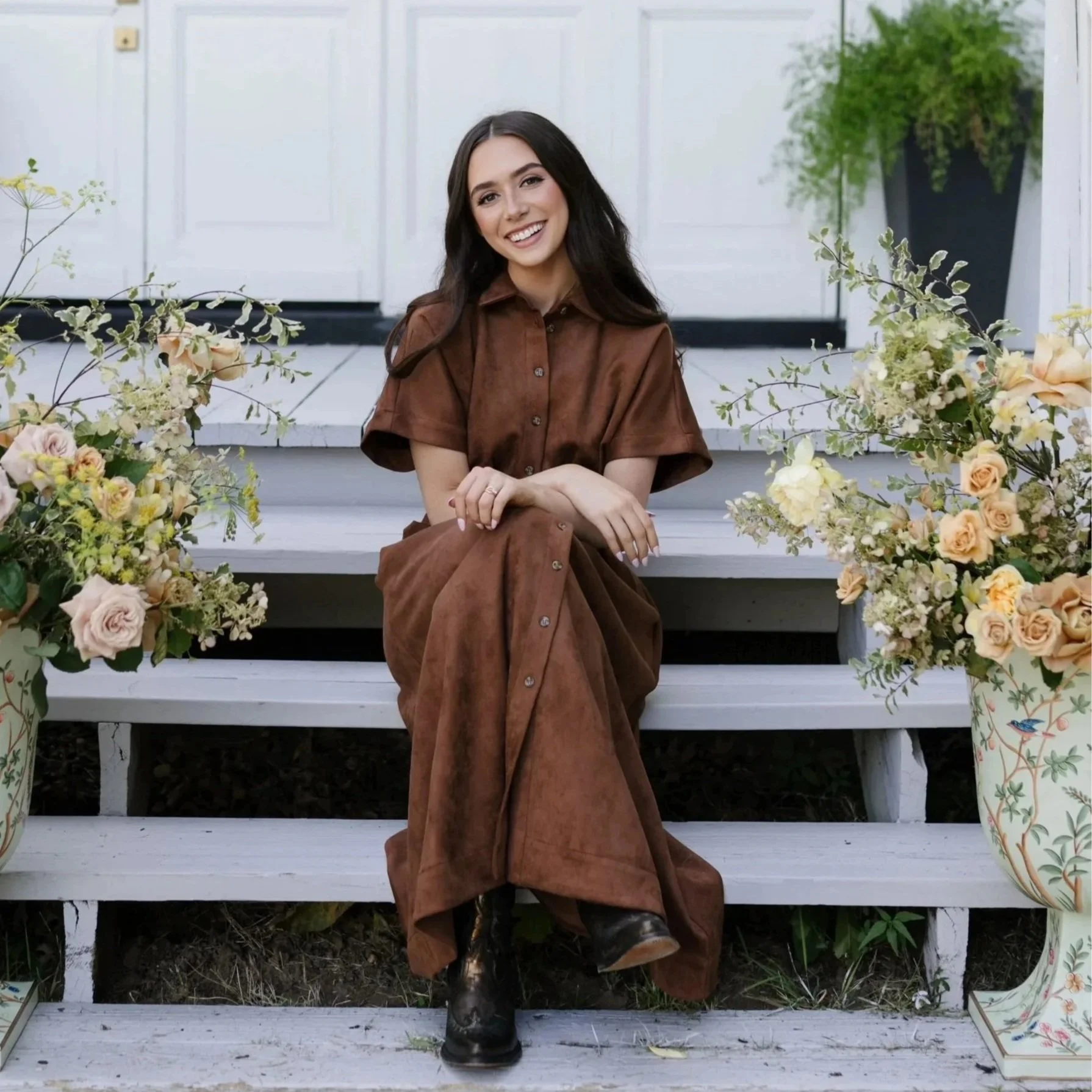 A young woman sitting on white wooden steps adorned with floral arrangements on either side, smiling with crossed arms, wearing a brown button-up dress and black boots, with a white house and green trees in the background.