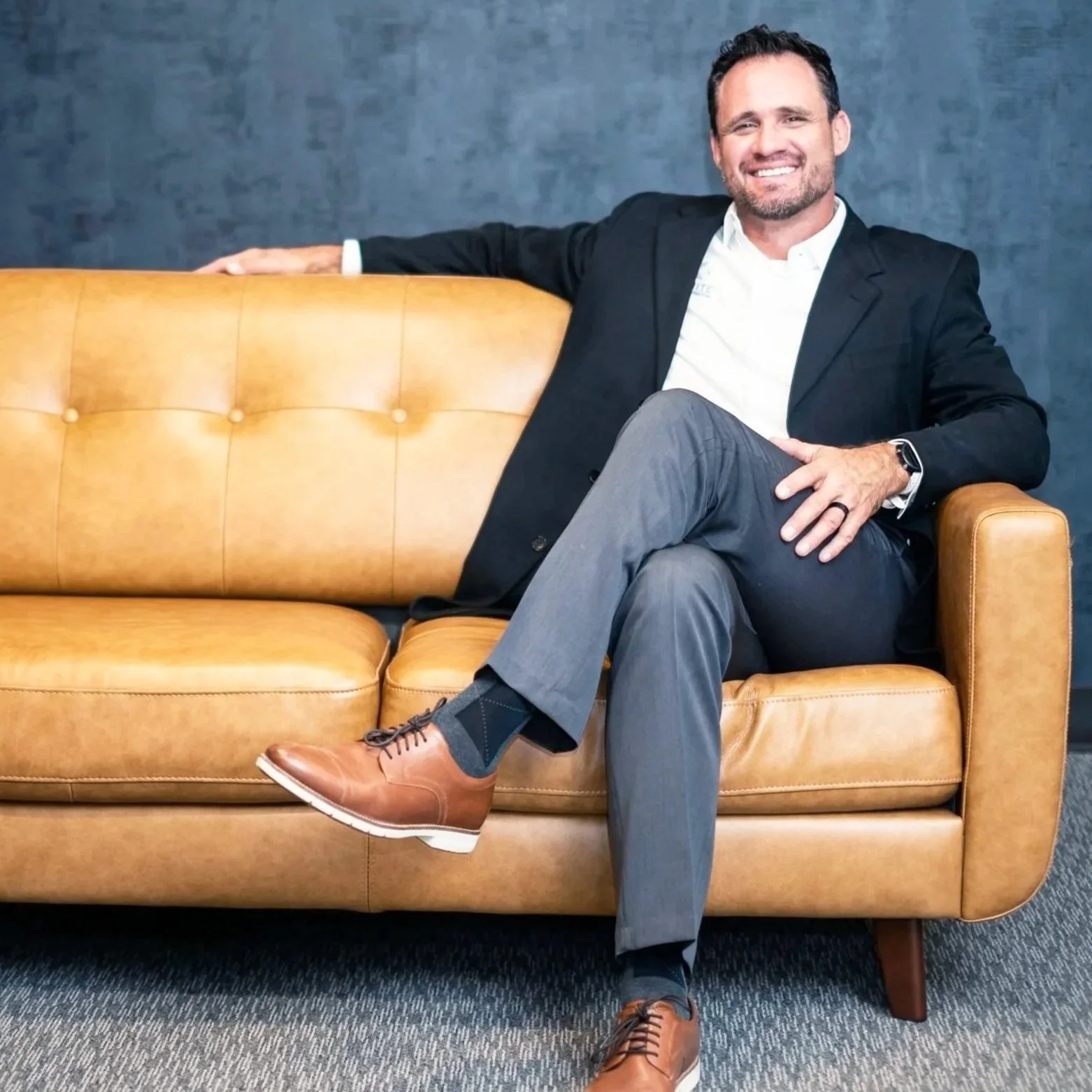 A man with dark hair, beard, and light skin, dressed in a black blazer, white shirt, gray pants, and brown shoes, sitting on a tan leather couch with a dark blue wall behind him, smiling at the camera with one arm stretched along the back of the couch and the other resting on his knee.