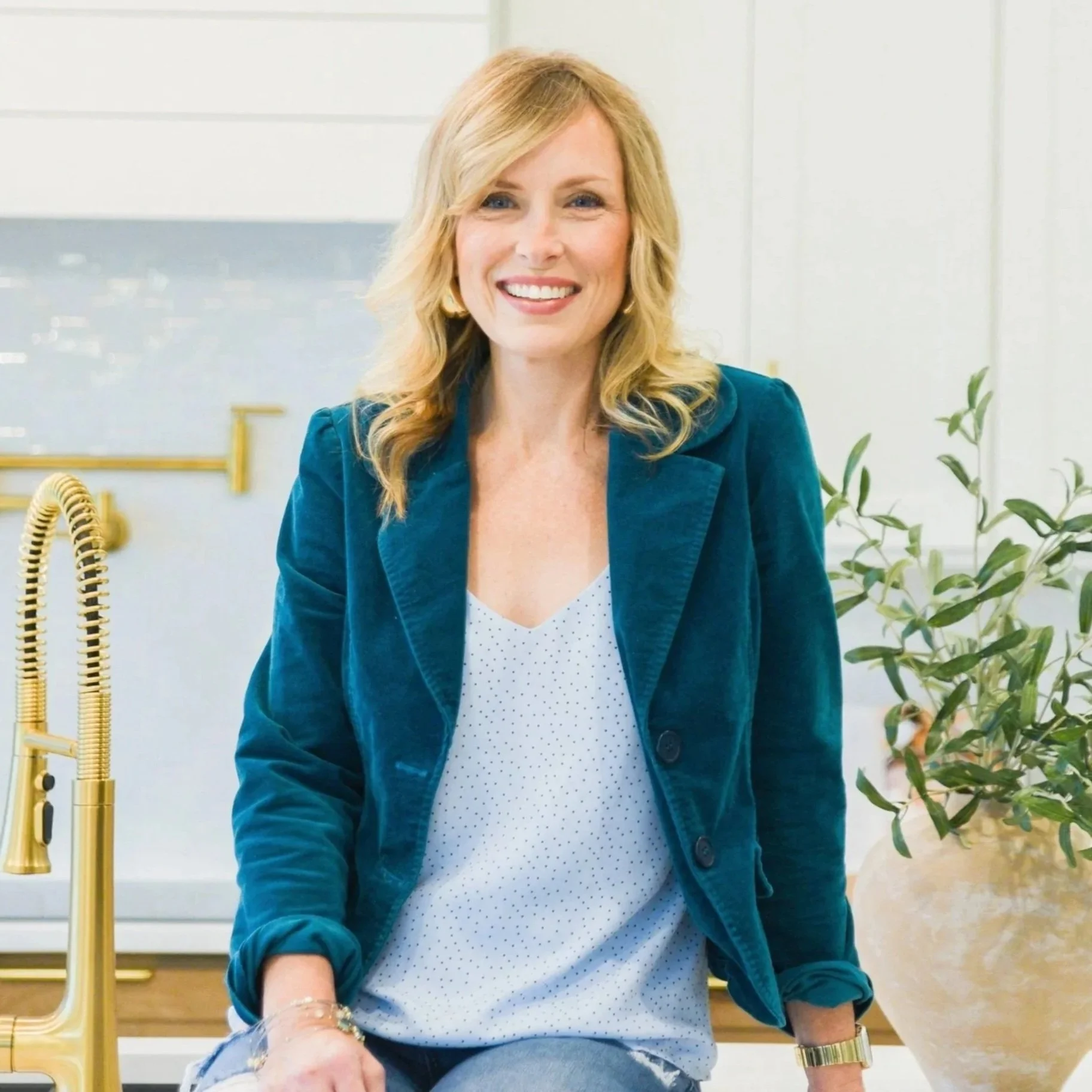 A woman with blonde hair and blue eyes smiling while sitting in a bright, modern room. She is wearing a teal jacket over a white polka dot blouse. In the background, there is a plant and a gold faucet with a gold soap dispenser nearby.
