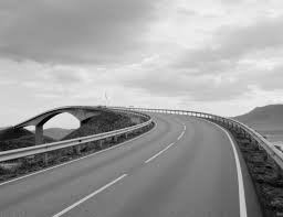 A curved highway overlooking water with hills in the distance, under cloudy skies.