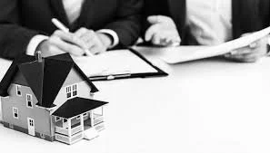 Two people in business attire working at a desk with a small model house in the foreground