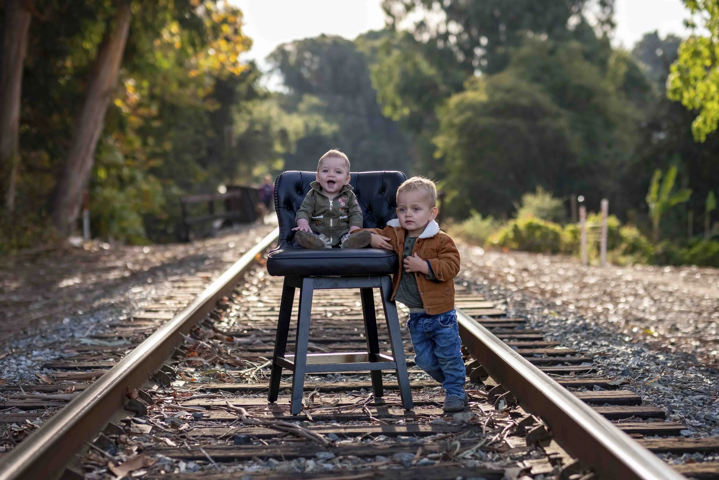 Two young children, one sitting on a chair on railway tracks and the other standing beside it, surrounded by trees with sunlight filtering through.