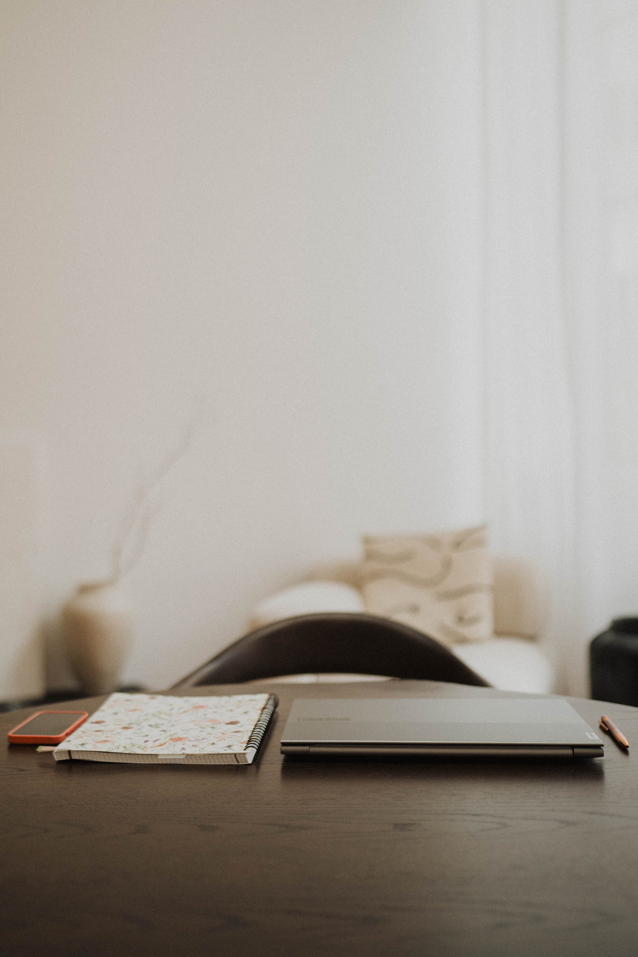 Een bureau met een open laptop, een notitieboek met een bloemenpatroon, een roze smartphone en een pen, met een bed en een vaas op de achtergrond.