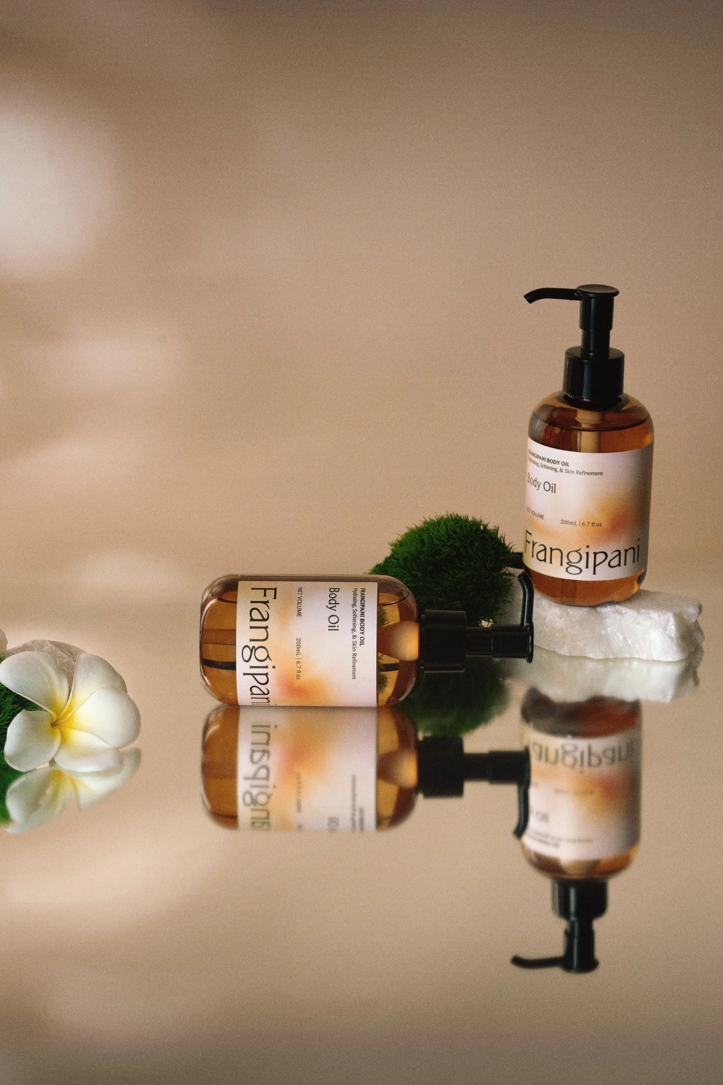 Two bottles of Frangipani body oil with a pump dispenser, a green moss ball, a white flower, and a white stone, all arranged on a reflective surface.