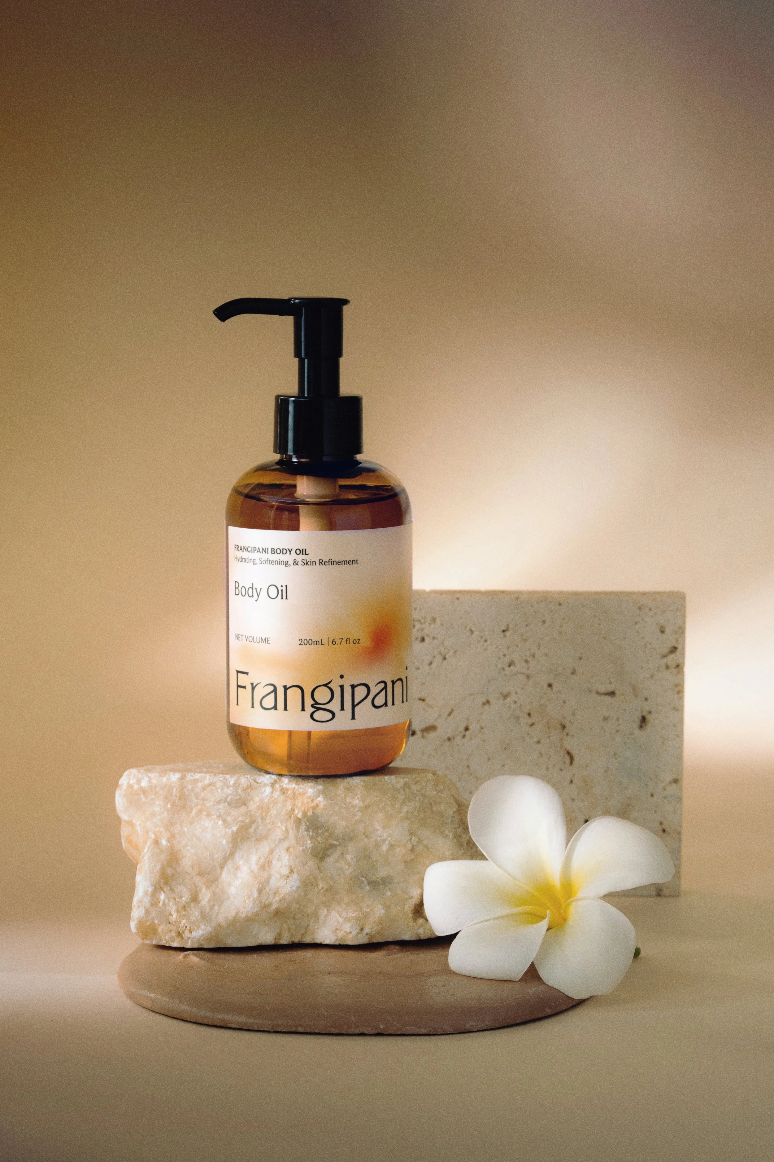 A bottle of Frangipani Body Oil with a pump dispenser, placed on a layered stone and wooden surface, accompanied by a white flower and a textured light-colored stone in the background.