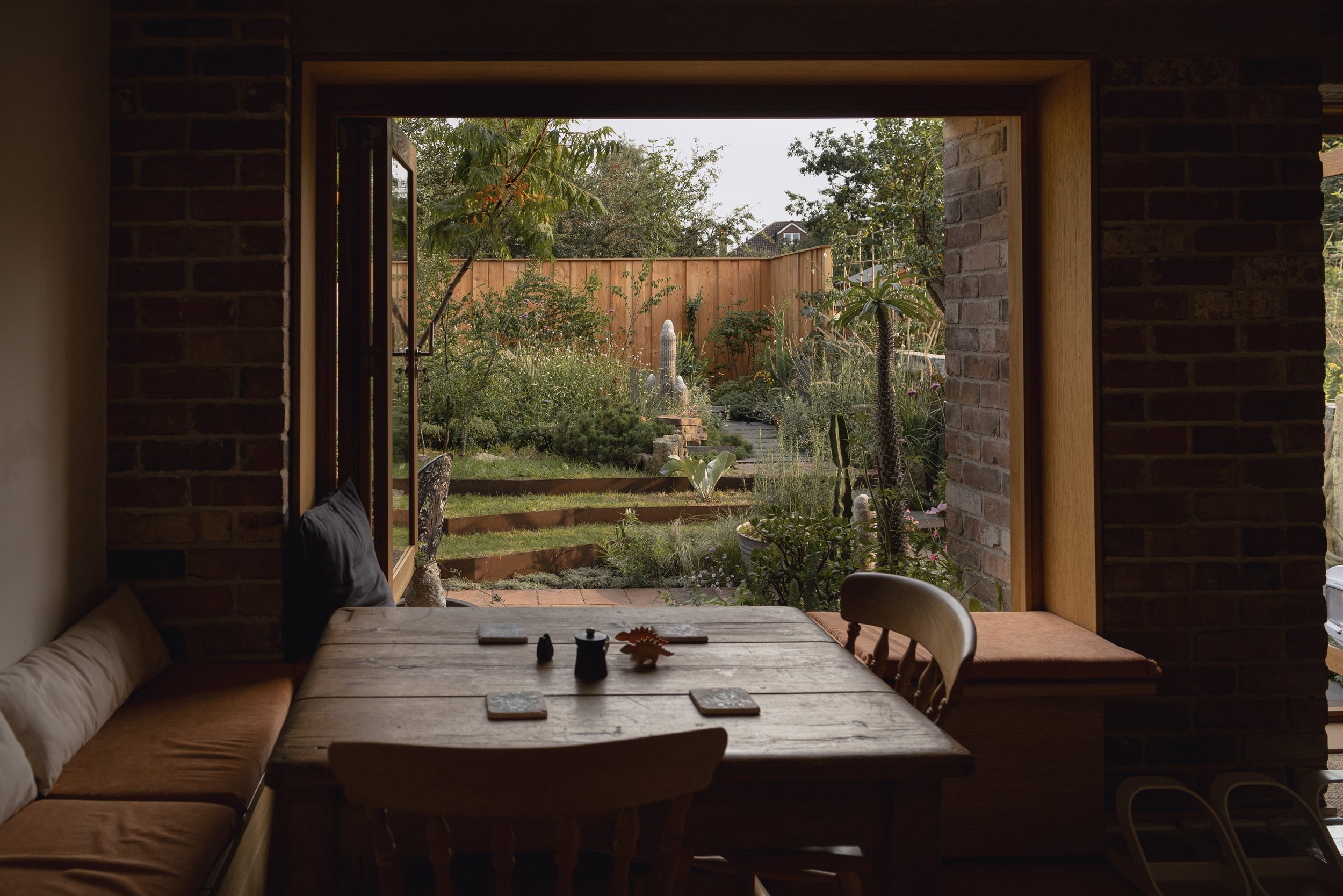 Cozy interior with wooden dining table and chairs, adjacent to an open door leading to a lush garden with plants and a wooden fence.