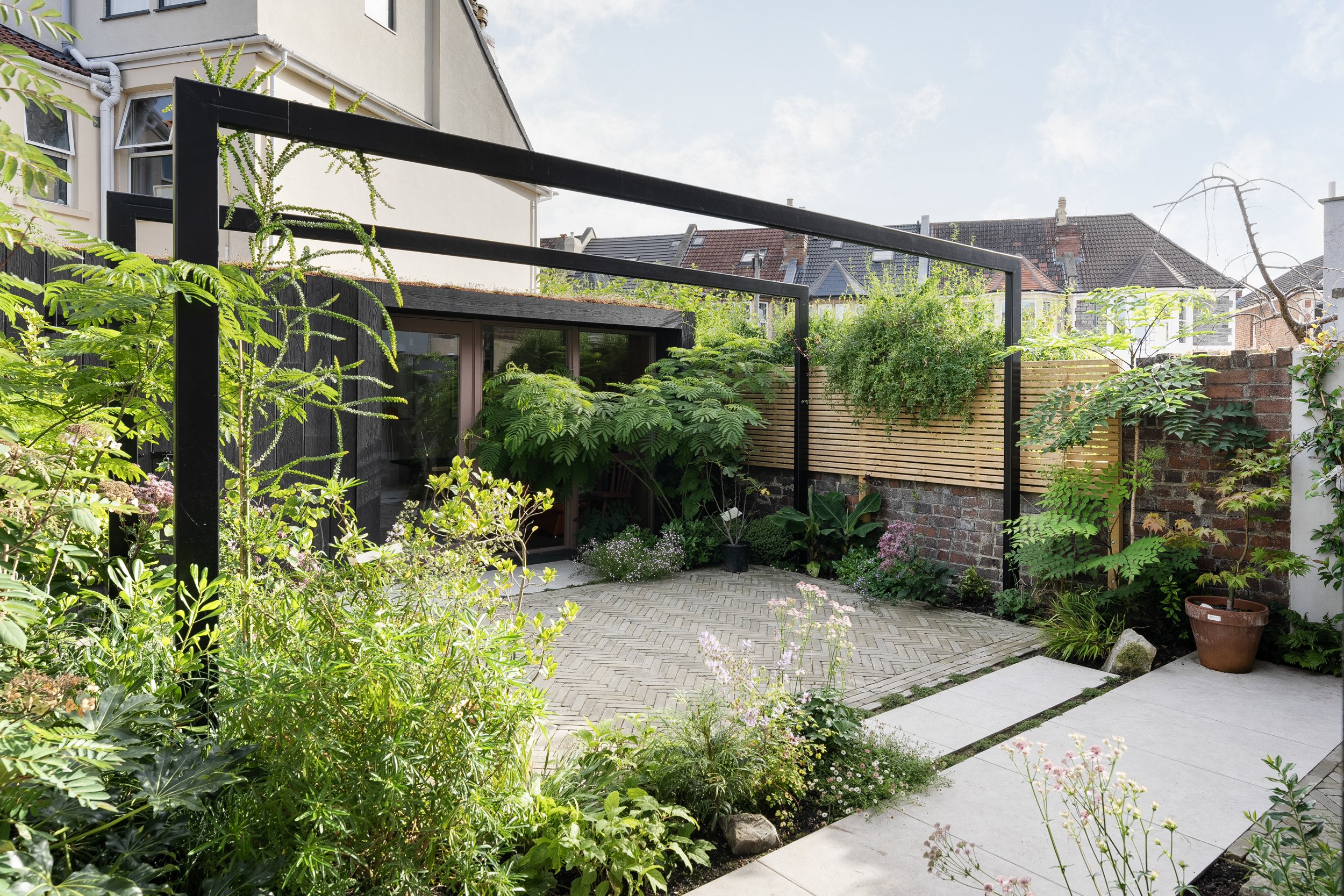 Modern garden courtyard with lush greenery, a brick and wooden fence, paving stones, and a large windowed building in the background.