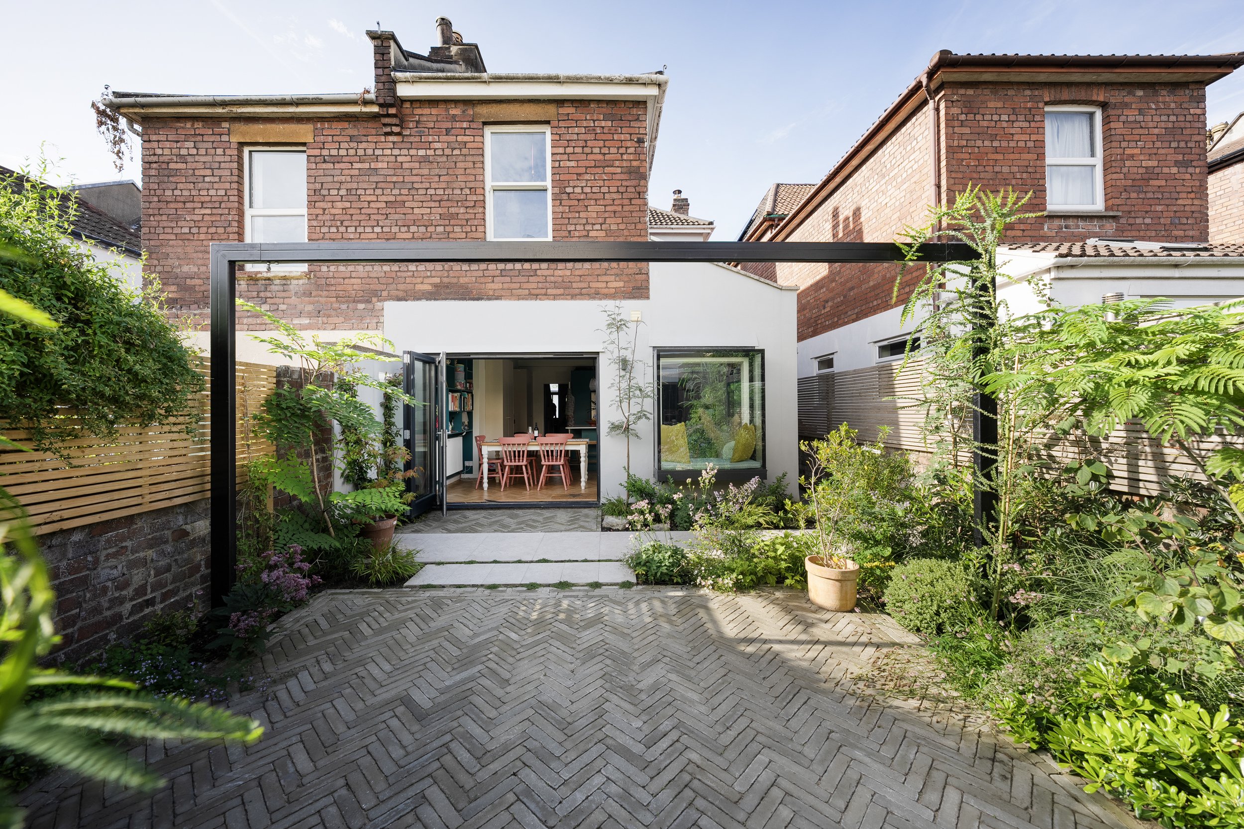 Backyard garden of a brick house with large glass doors and a patio, surrounded by lush greenery and potted plants.