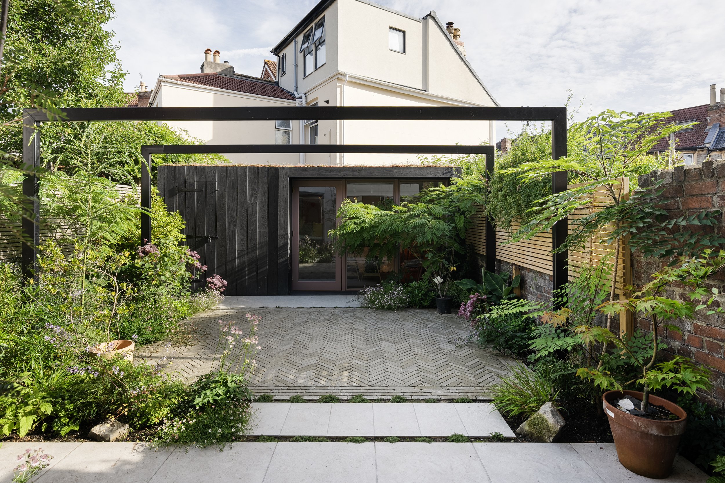 Modern backyard with a small structure, surrounded by lush greenery and plants, herringbone paving stones, and privacy fencing.