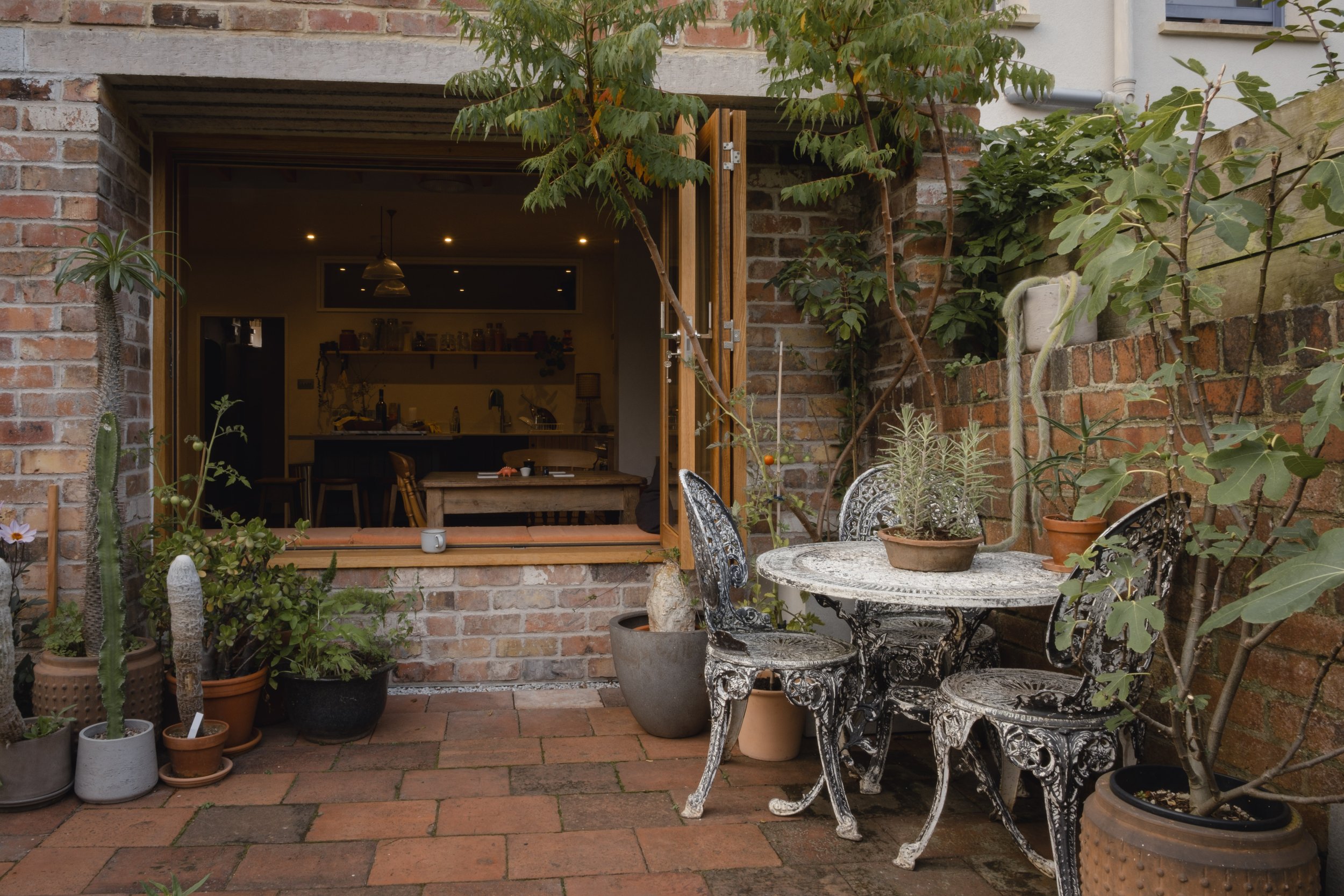 Outdoor patio with decorative metal table and chairs, surrounded by potted plants and cacti, adjacent to a brick house with an open sliding door leading to a kitchen area.