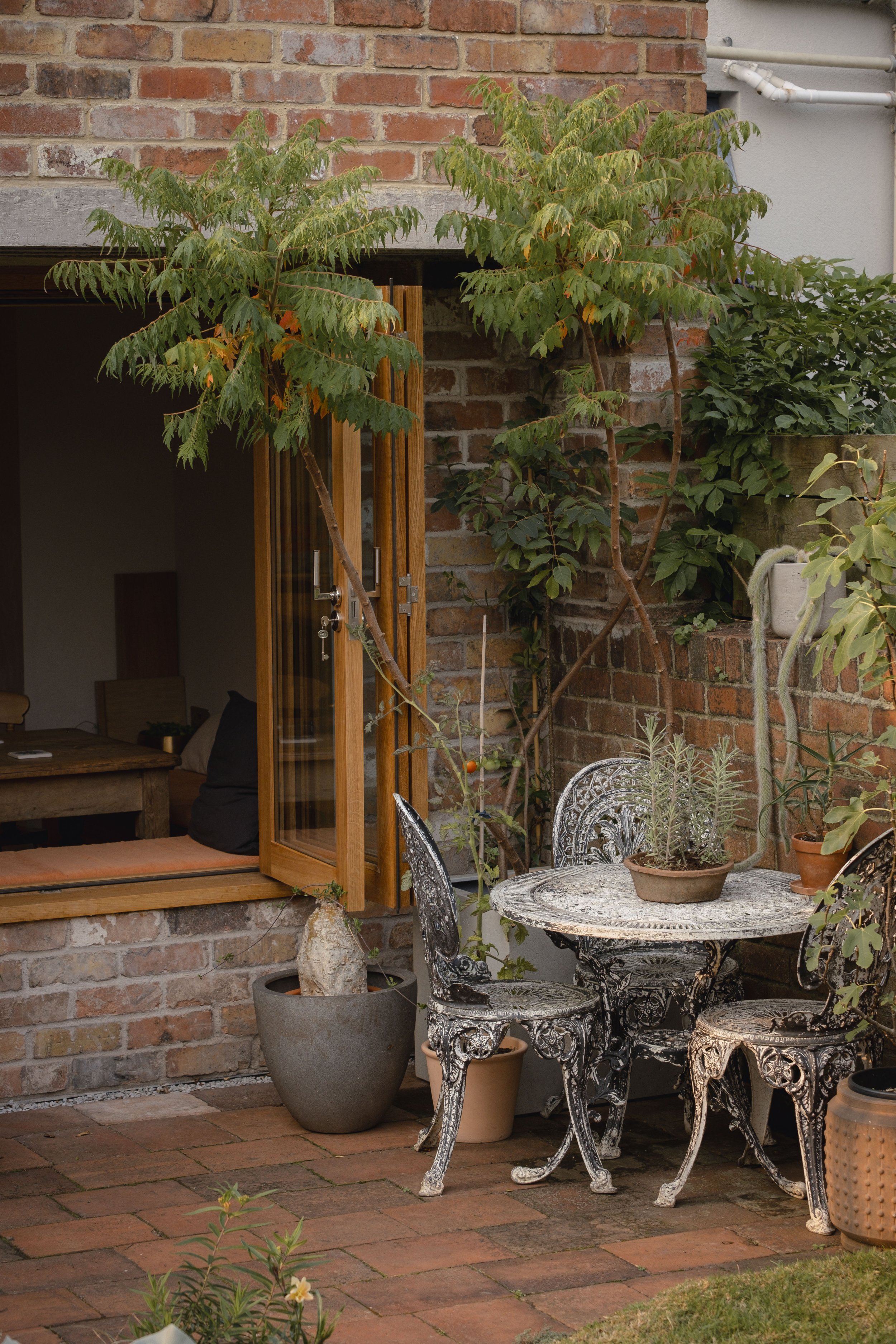 Cozy outdoor patio with intricately designed metal table and chairs, potted plants, open wooden door, brick wall backdrop, and a small tree.