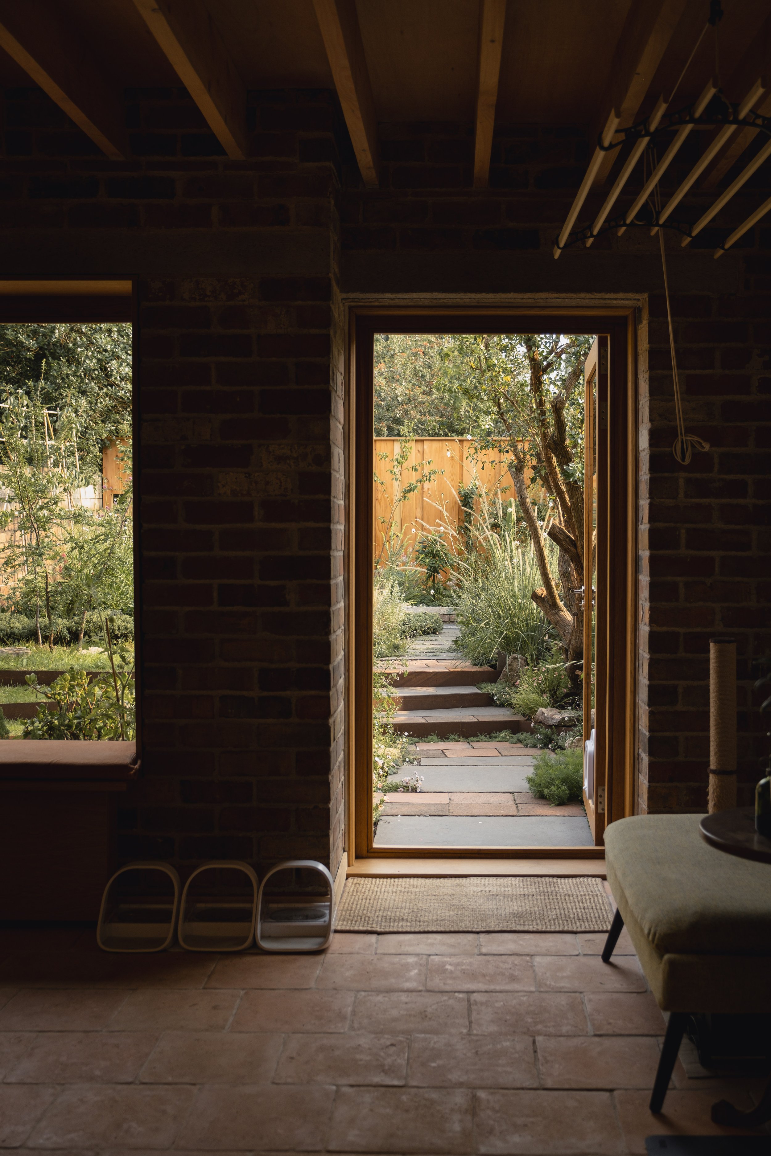 Interior view of a brick-walled room with an open door leading to a garden path, surrounded by lush greenery and a wooden fence. Sunlight illuminates the garden path, shaded by trees.
