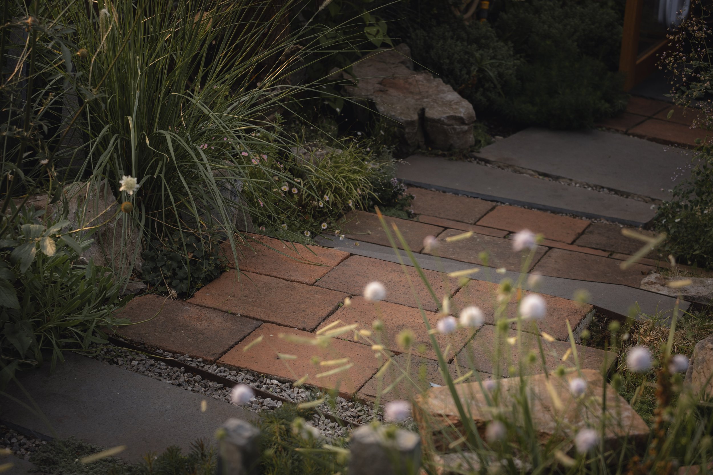 Garden pathway with bricks and stones surrounded by lush greenery and wildflowers.
