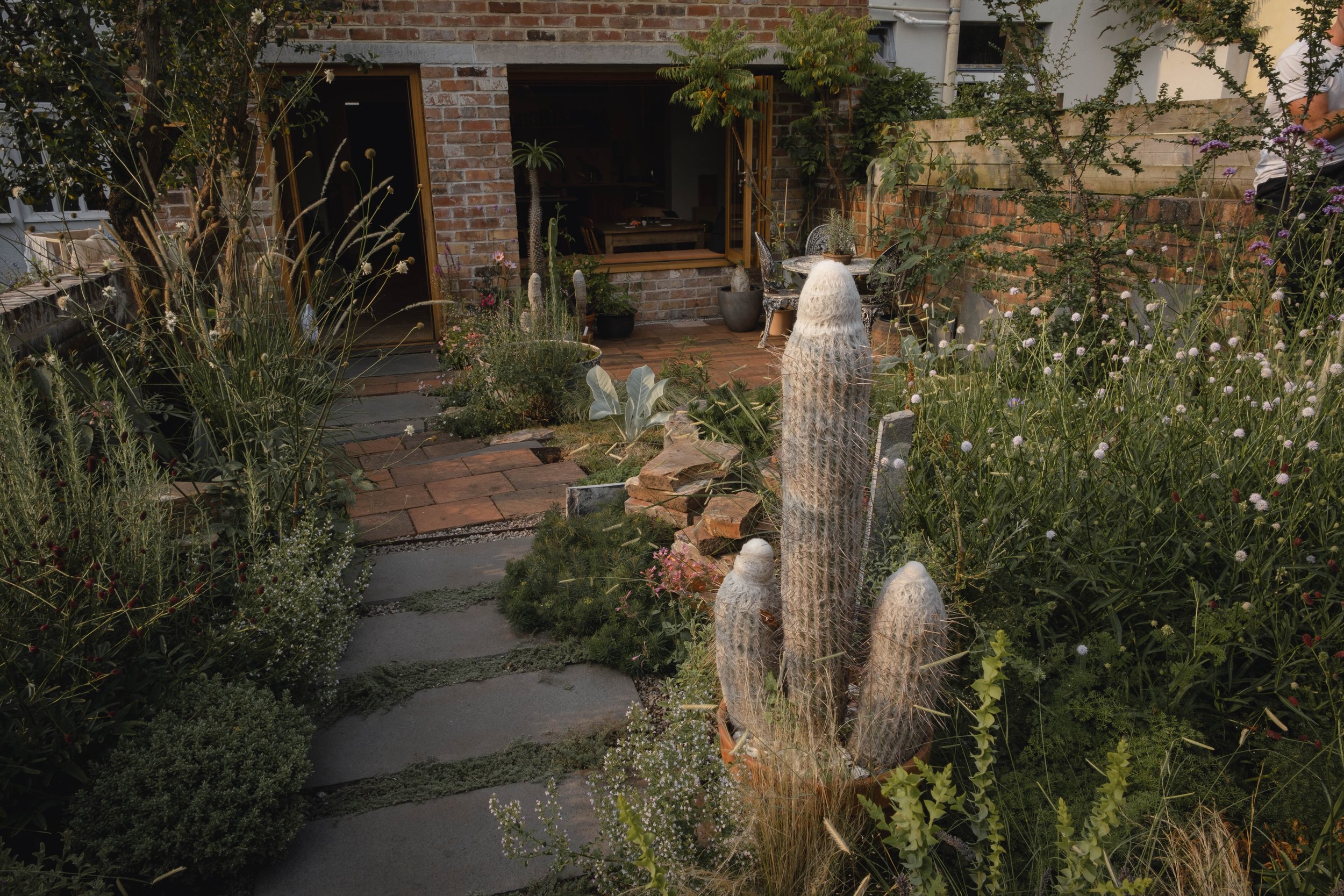 A garden with various plants, including tall cacti wrapped in protective netting, and a brick walkway leading to a building with open wooden doors. There's outdoor seating and greenery throughout the space, creating a lush and inviting atmosphere.