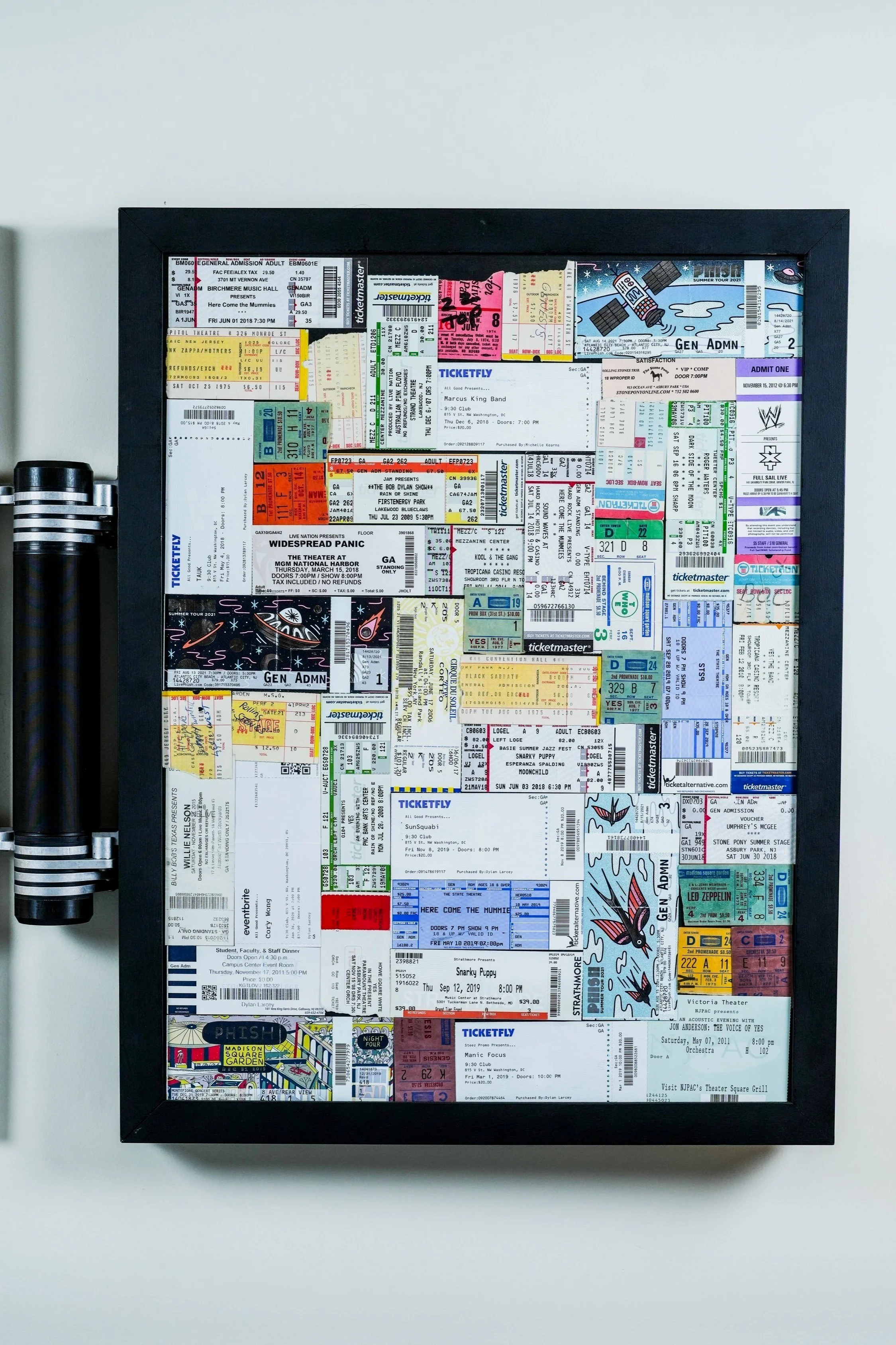 A framed collage of various colorful concert tickets displayed on a light gray wall.
