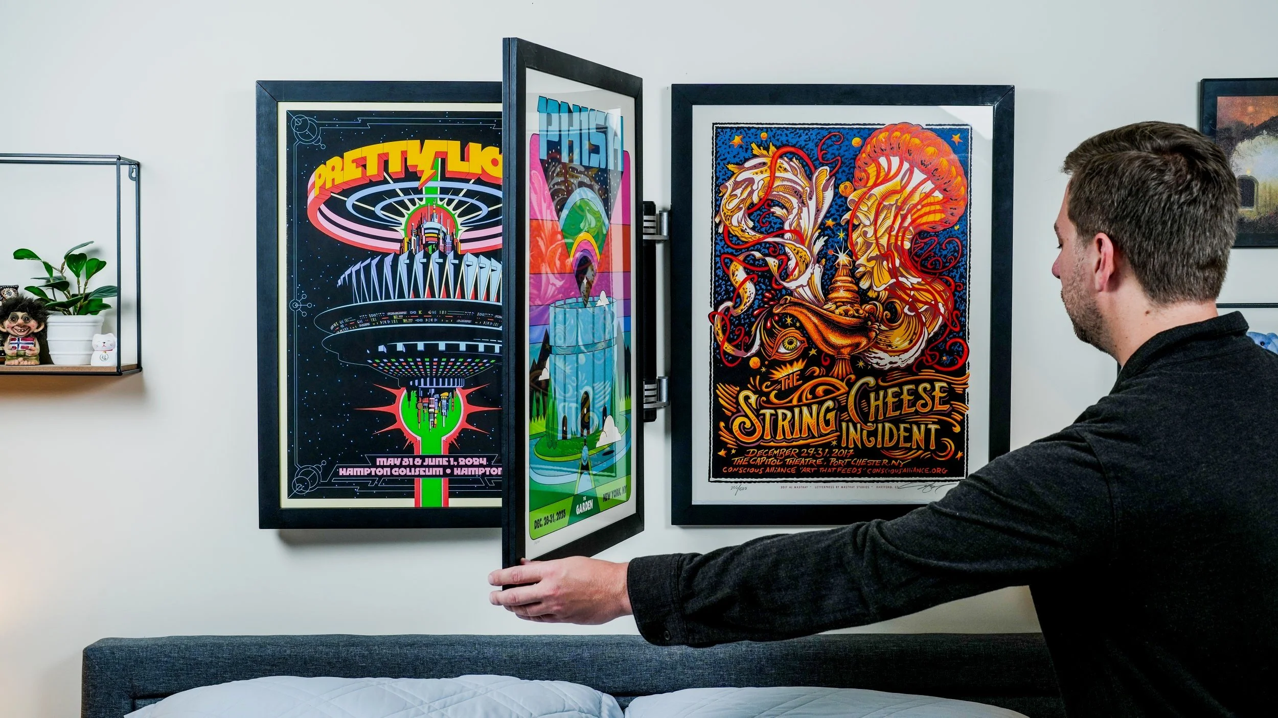 A man with gray hair and beard adjusting a framed concert poster on a white wall in a room with minimal bedroom decor.
