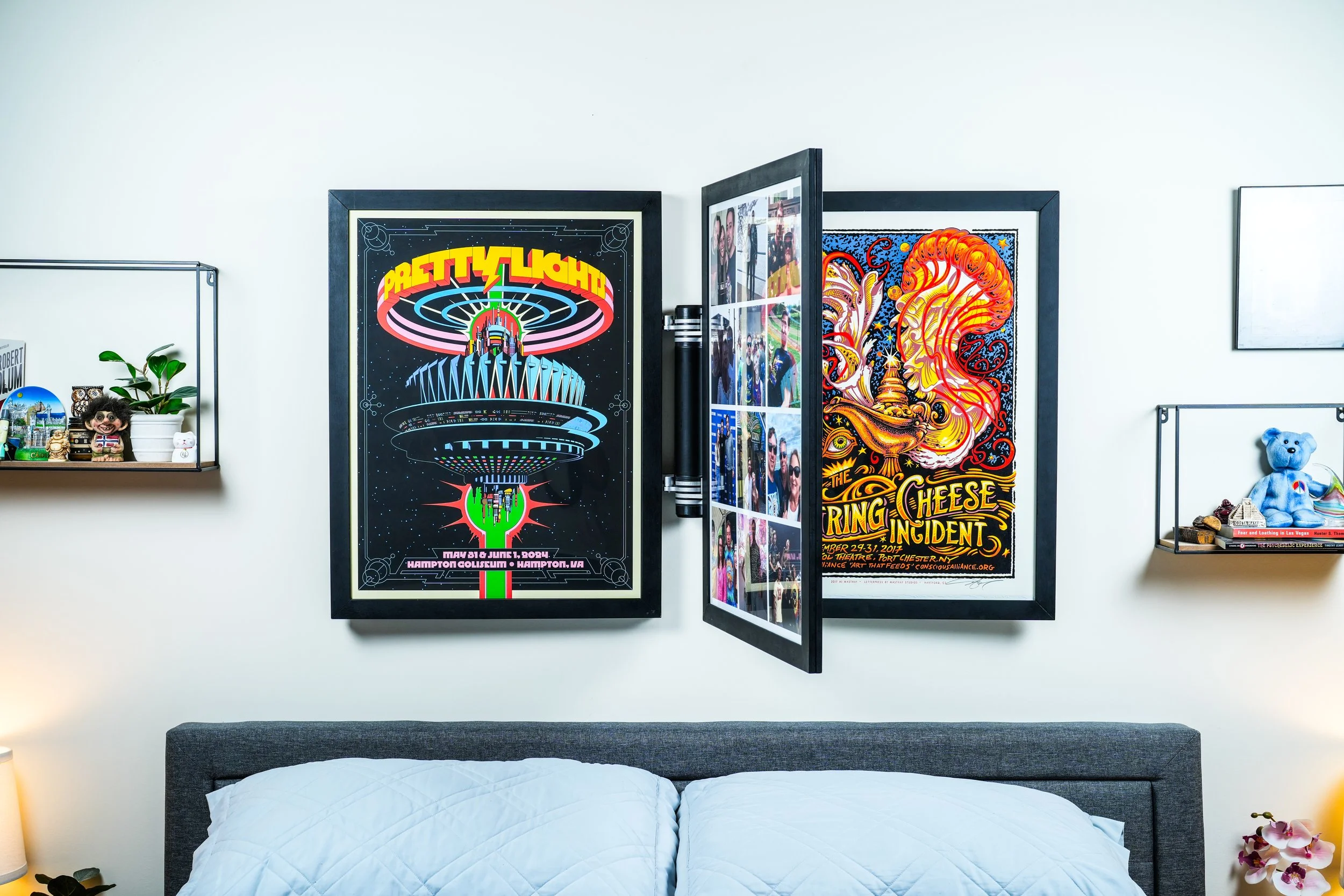 View of a bedroom wall decorated with posters; one poster featuring a colorful roller coaster with the word "Pretty Lips" and event details, and another showcasing a vibrant, artistic design with the text "The Spring Cheese Incident"; a shelf on the left with plants, figurines, and souvenirs, and a bedroom with a bed covered in a white quilt at the bottom.
