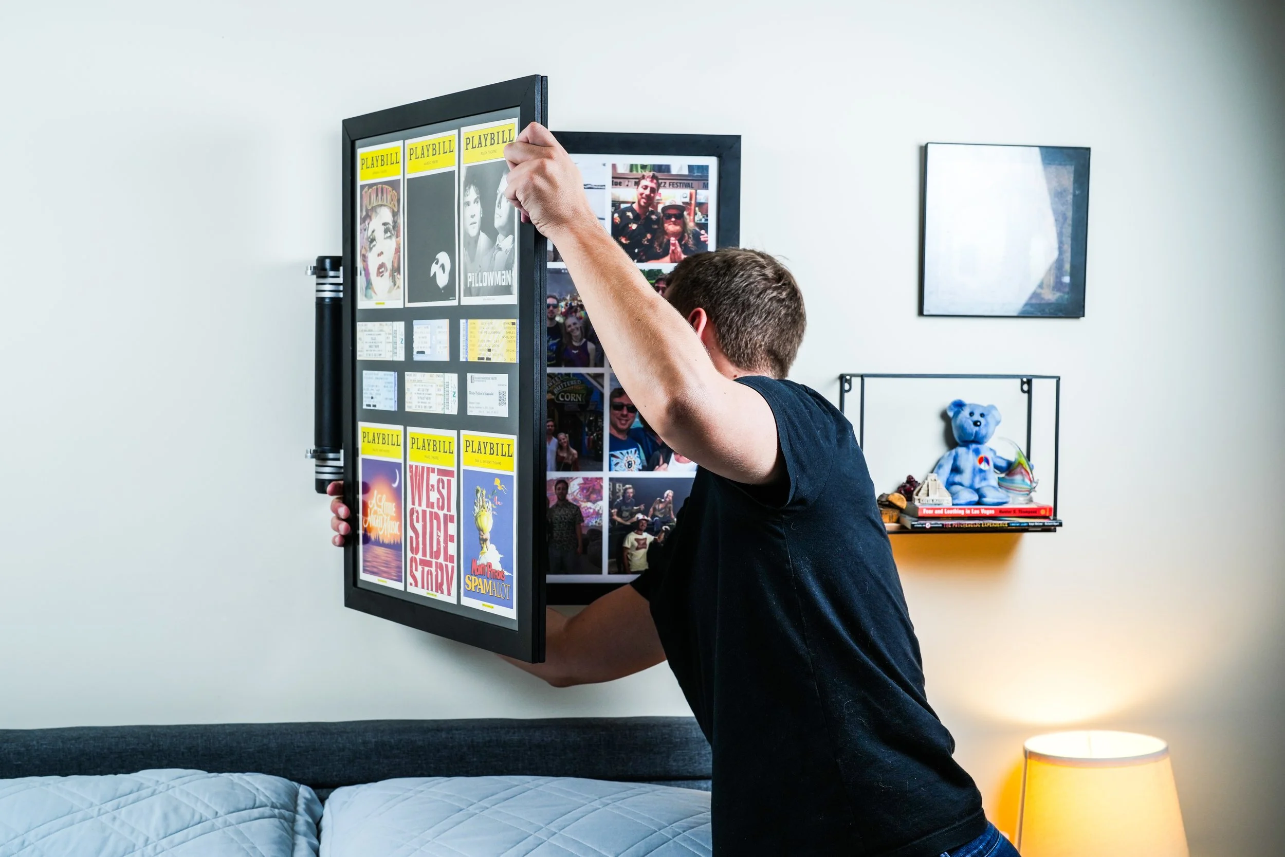 A man is hanging a framed display of theater playbills on a wall in a bedroom, partially blocking a bed and a small bookshelf with books and a teddy bear.