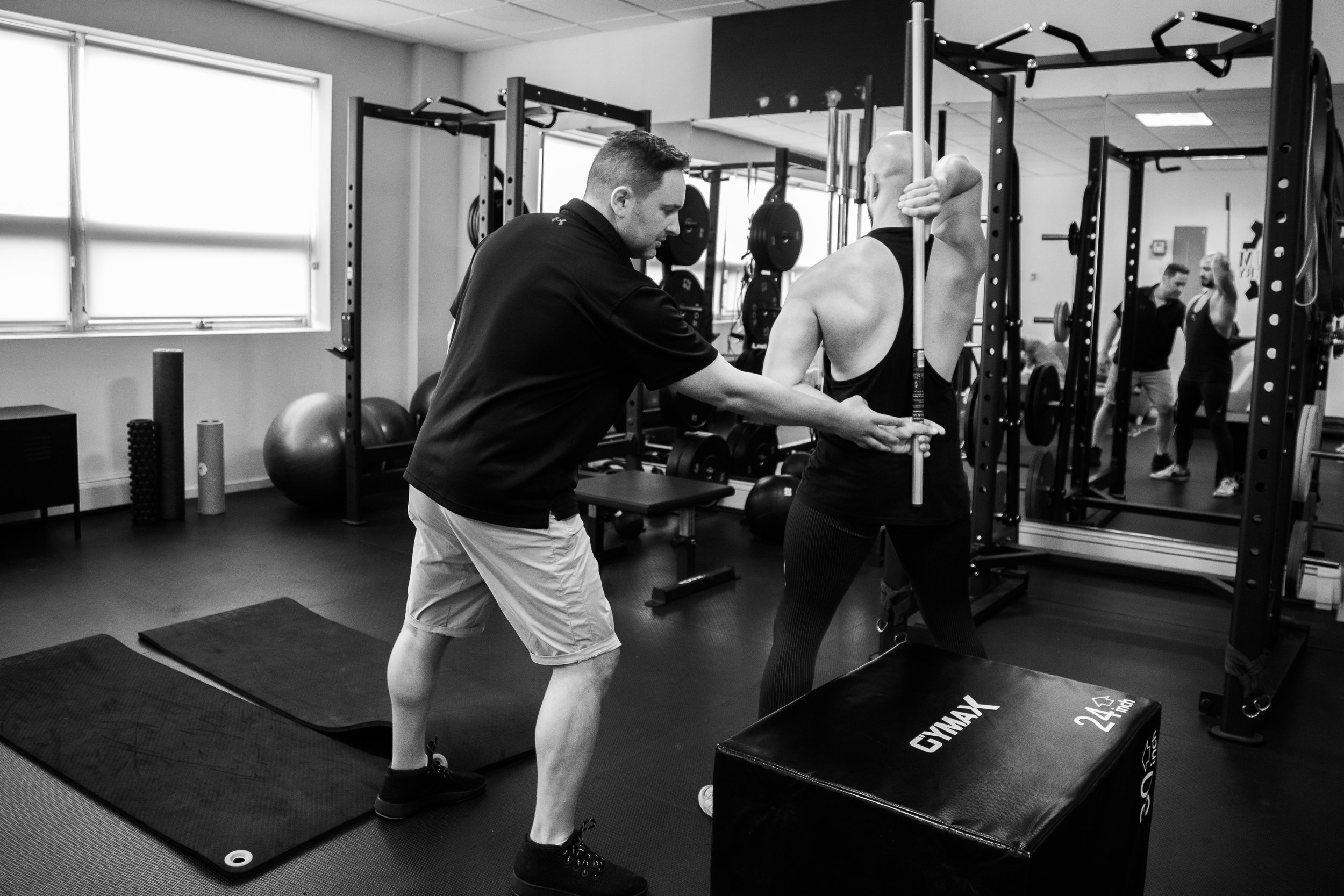 A personal trainer assisting a bald man with muscular back during a workout in a gym, with other people in the background.