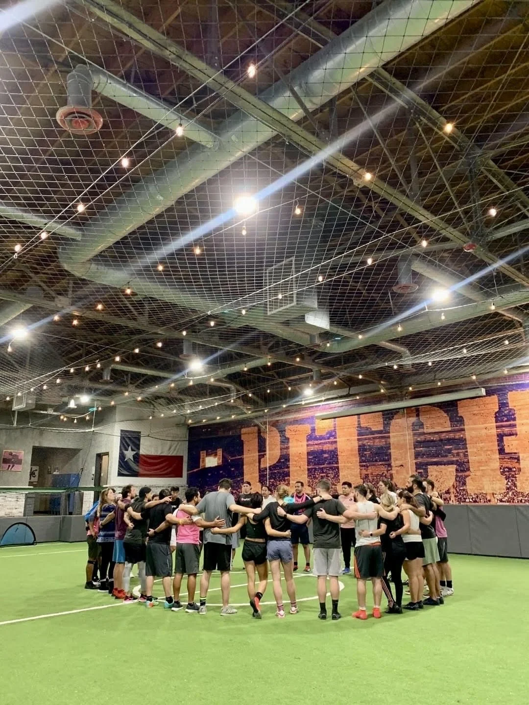 Group of sphere™ people playing soccer on a sunny outdoor field, wearing pink training vests and black shorts, near a banner with 'sphere' logo.