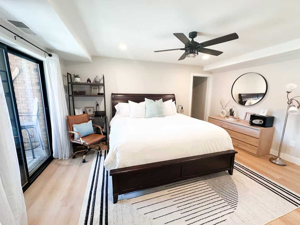 Bedroom with a large bed, black ceiling fan, sliding glass door with white curtains, wooden dresser with mirror, black shelving unit, brown chair, and beige rug.