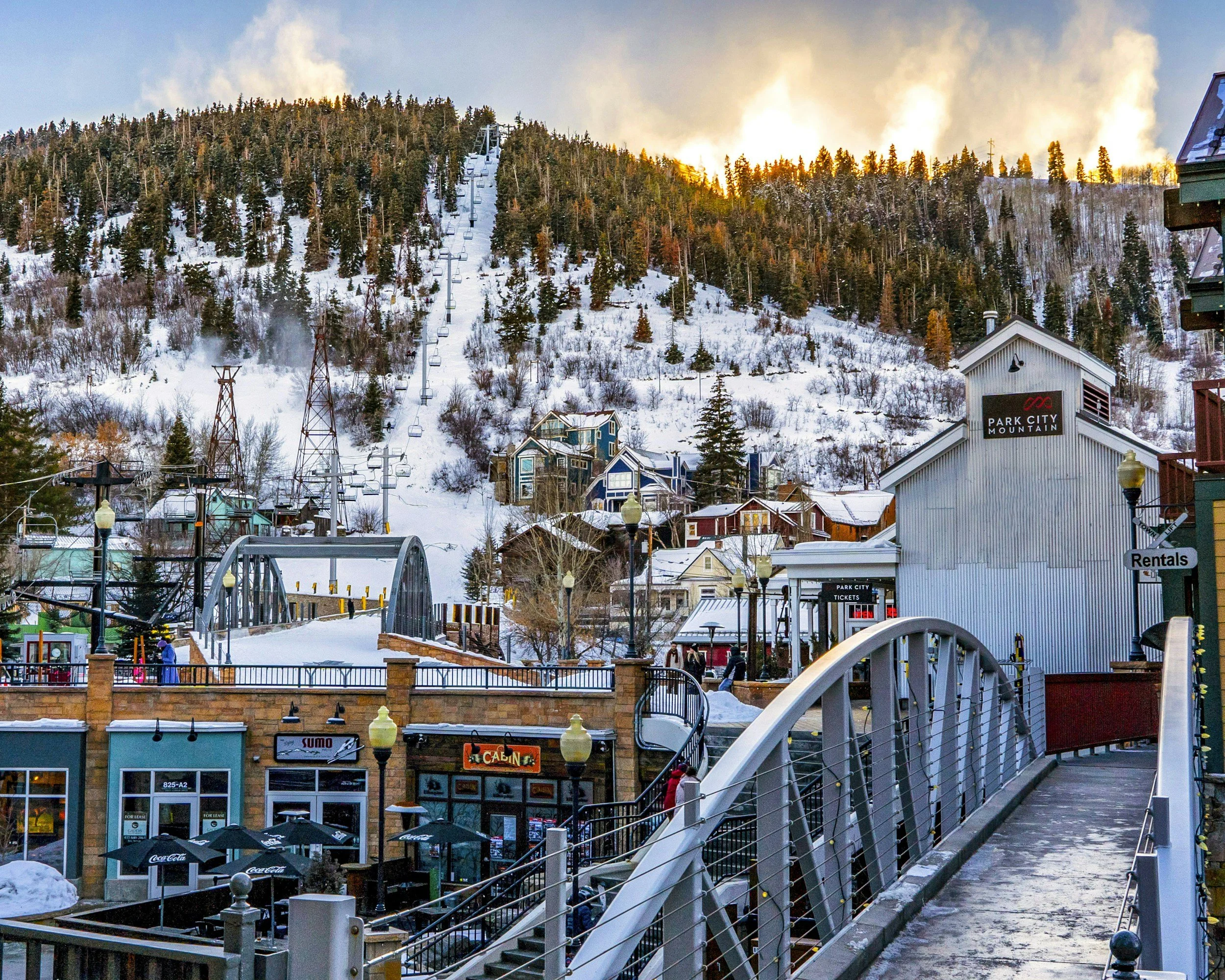 Snow-covered buildings at the base of a mountain with ski lifts and trees during sunset