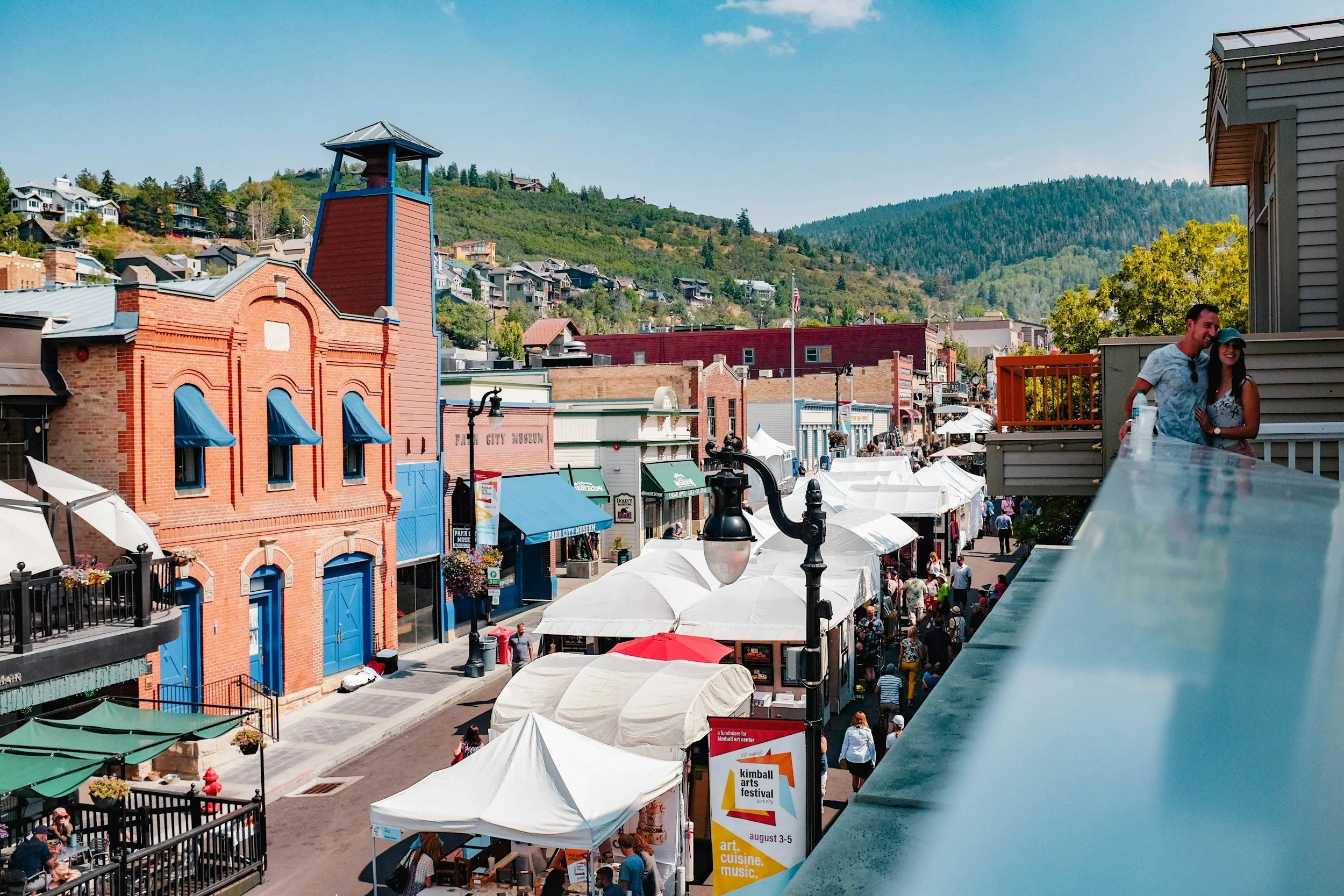 View of a bustling street fair with tents and umbrellas, colorful buildings with blue awnings, people walking and shopping, and a backdrop of green hills under a clear blue sky.