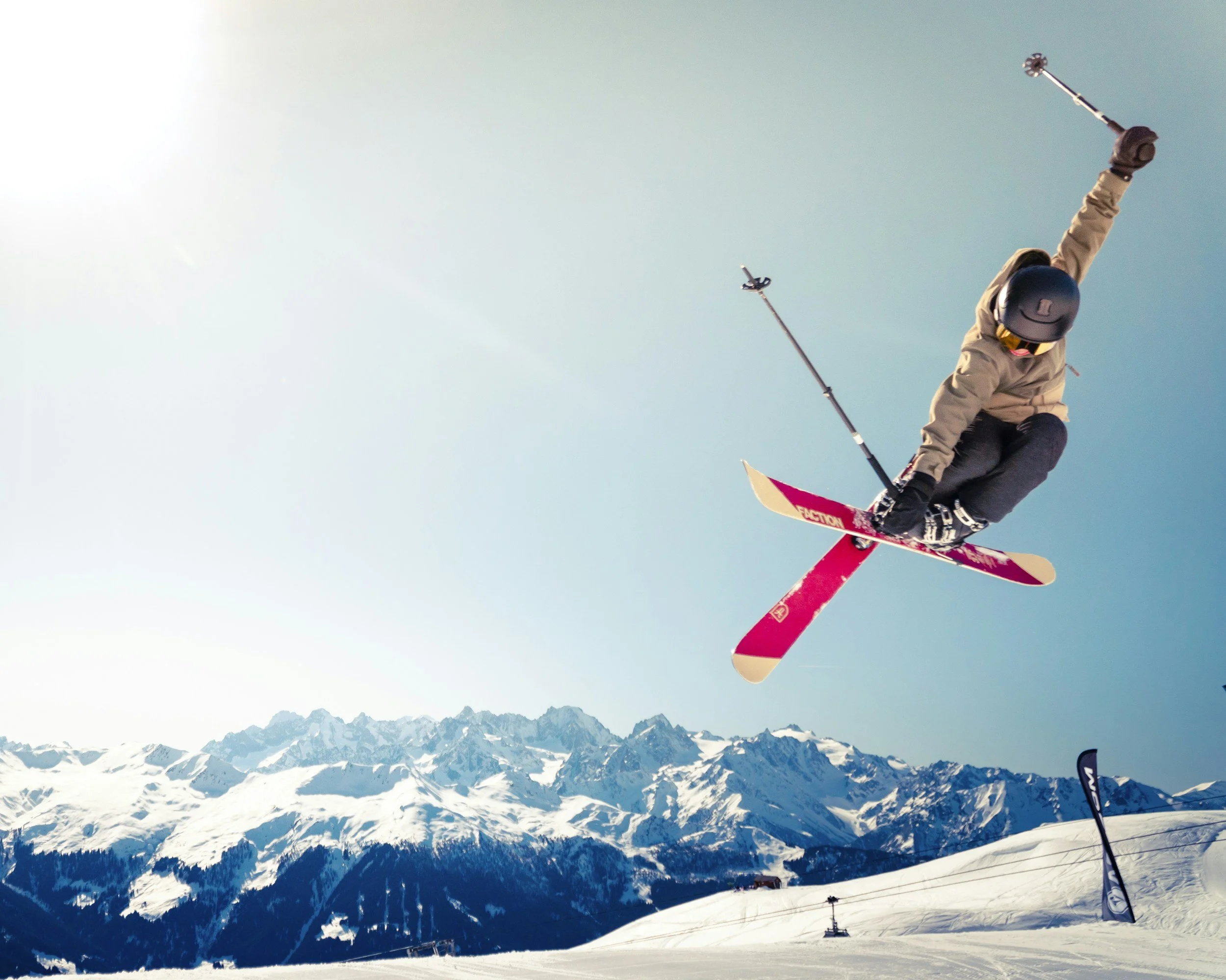 A person skiing in the mountains under a blue sky, with snow-covered peaks in the background.