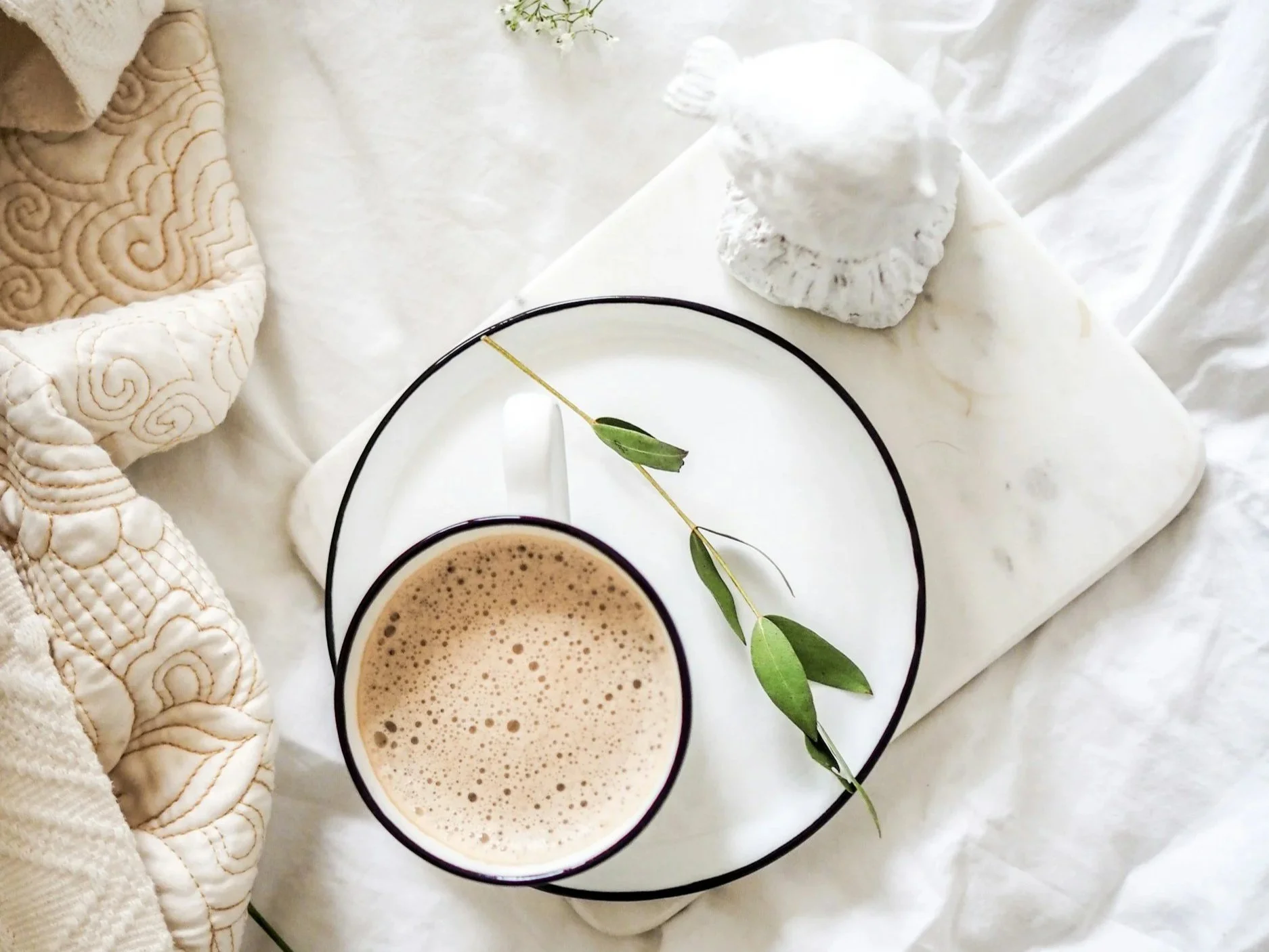 A cup of frothy coffee or hot chocolate on a white plate with green leaves, placed on a cream-colored fabric background with a beige quilted blanket and a white decorative stone sculpture nearby.