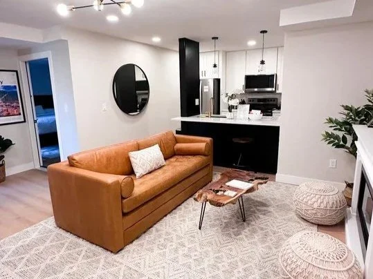 Living room with a tan leather sofa, a small coffee table, a round mirror on the wall, and a kitchen area with black cabinets and stainless steel appliances in the background.