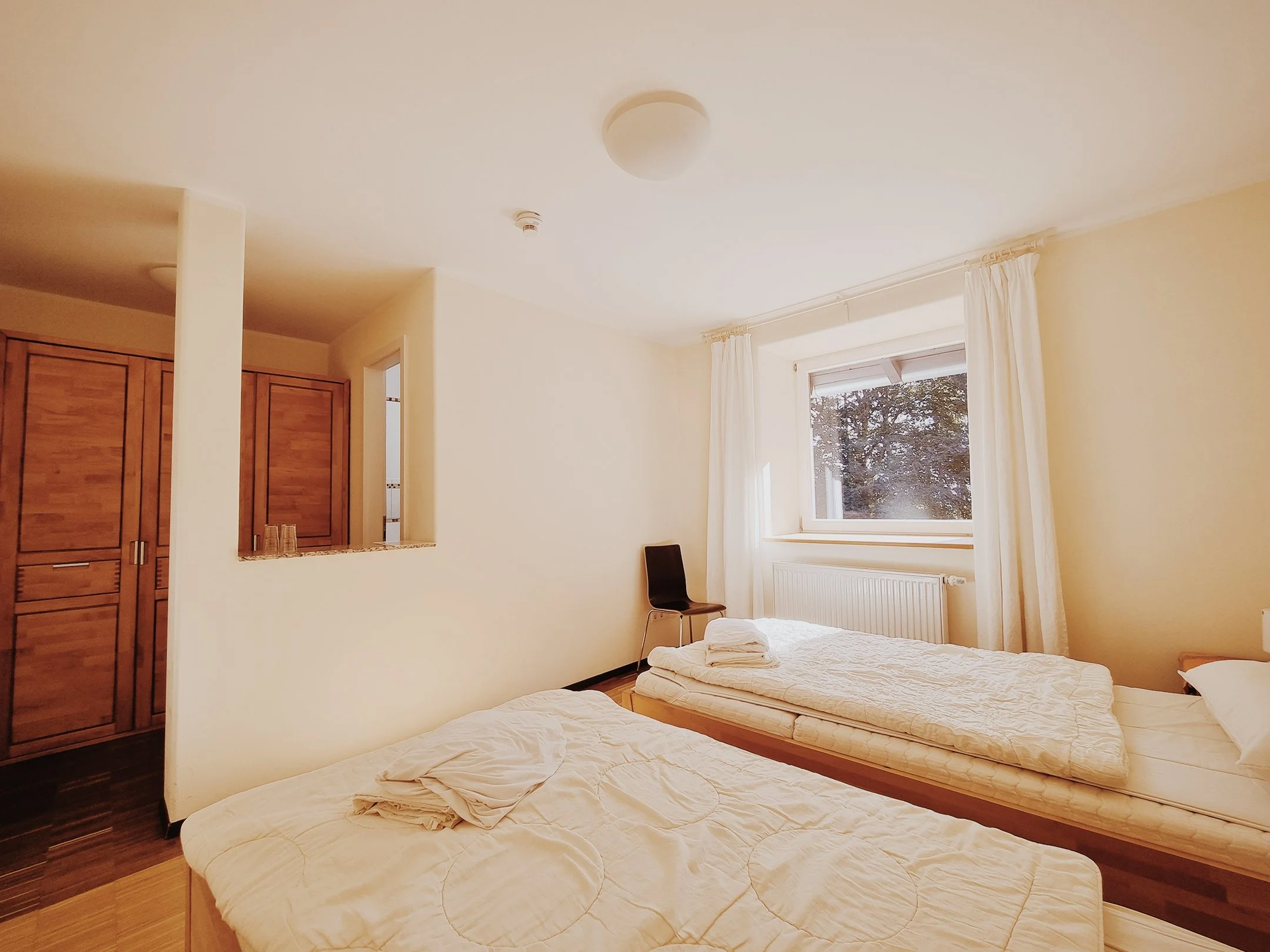 Zimmer mit zwei Betten, Holzschrank und Fenster mit weißen Vorhängen