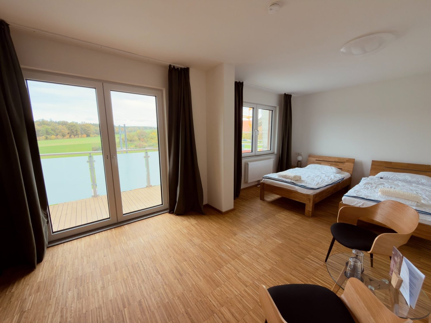 Premium-Zimmer mit zwei Holzbetten, Holzboden und bodentiefer Fenstertür zum Balkon mit Blick über die Felder